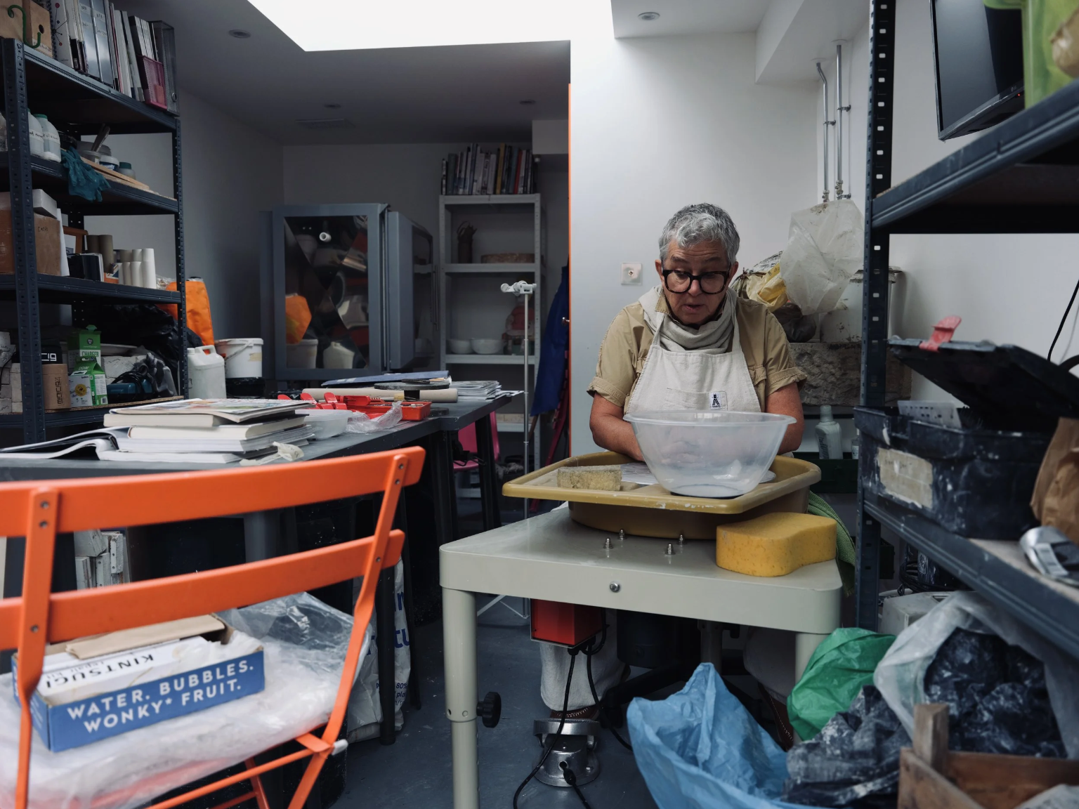 An older person with gray hair and glasses, wearing a beige apron, working at a table in a cluttered workshop or storage room. They are focused on a task with various containers and tools around them.