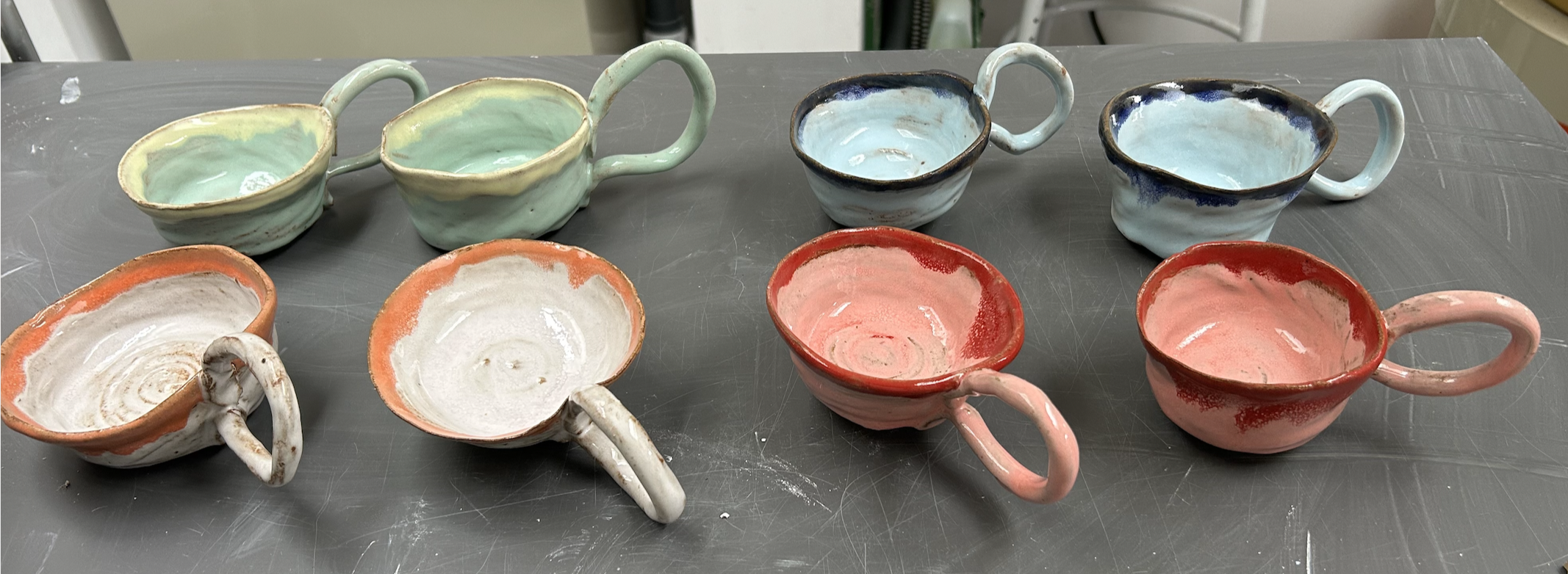 Colorful handmade ceramic cups with handles, arranged on a gray surface, in shades of green, blue, white, orange, and pink.