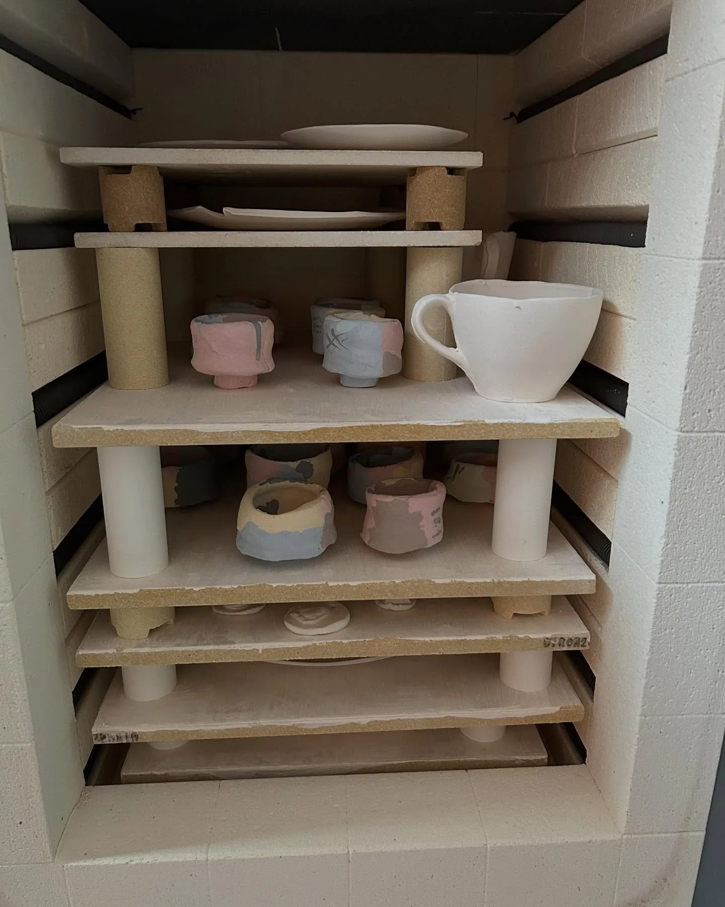 A kiln shelf with various unfinished ceramic cups and bowls in a kiln.