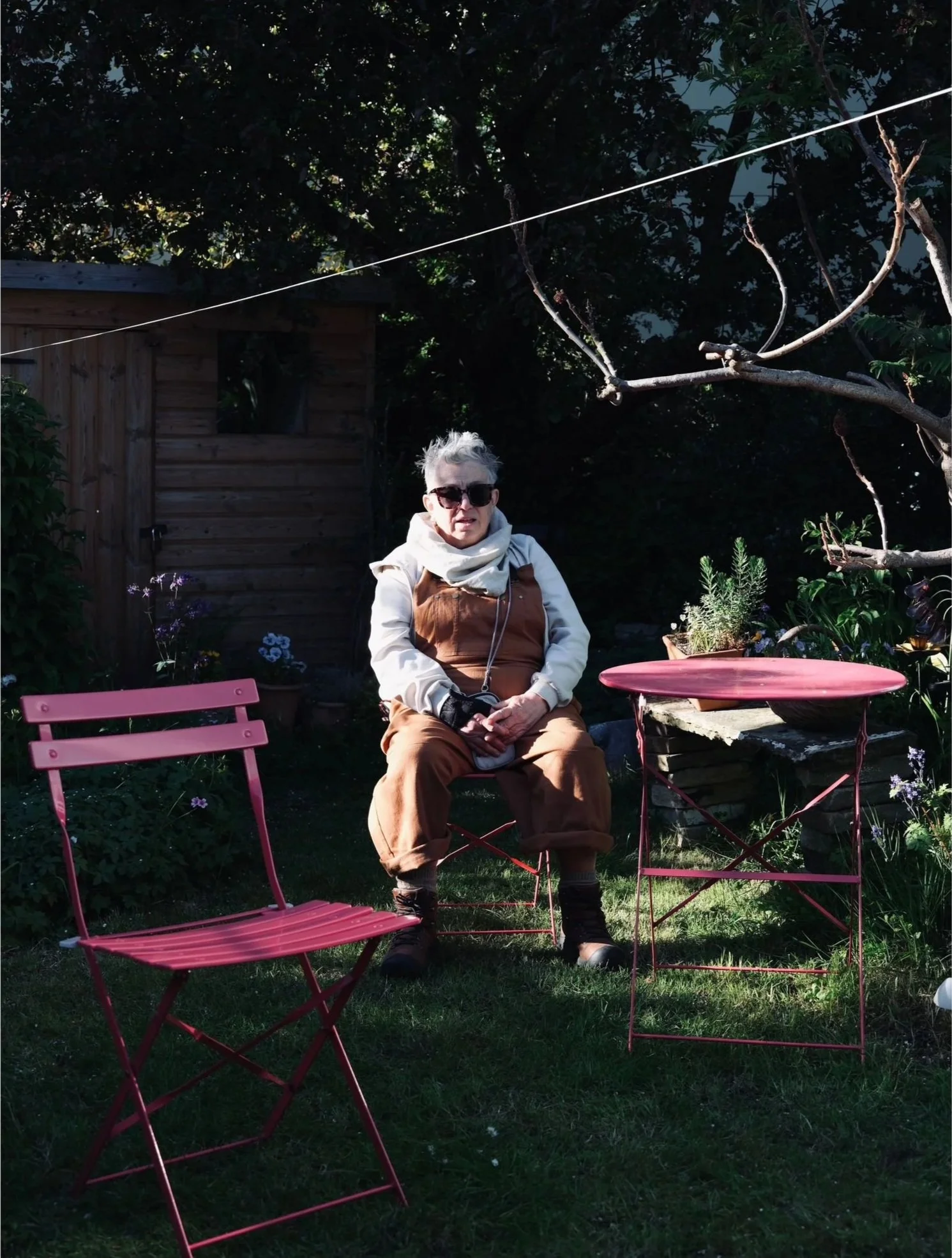 An elderly woman with gray hair, wearing sunglasses, a beige jacket with a hood, and brown pants, is sitting outdoors in a garden. She is surrounded by pink metal chairs and a pink metal table. Behind her is a small wooden shed, and there are trees and plants in the background. The woman appears relaxed and is looking toward the camera.