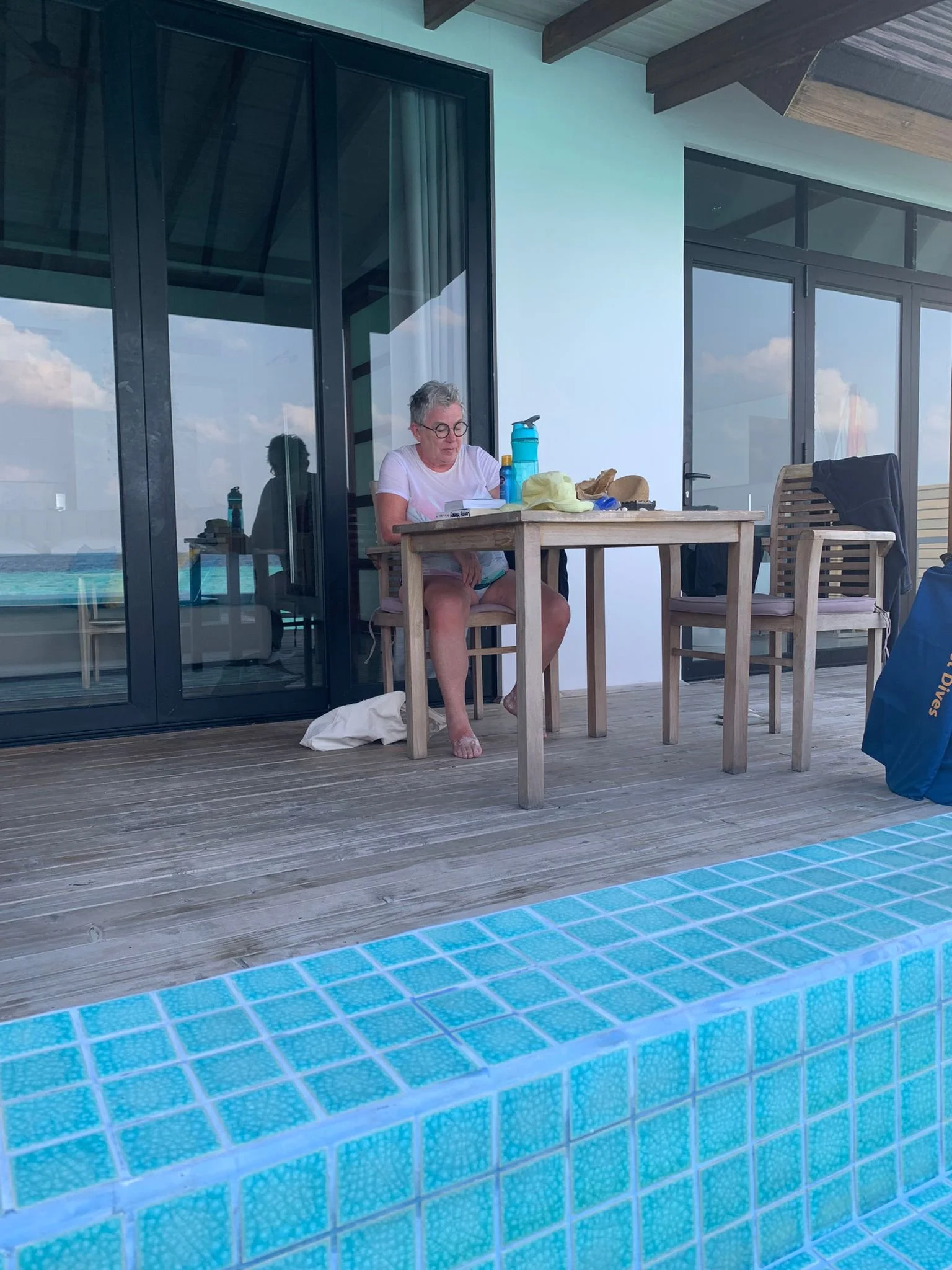 Woman sitting at an outdoor table near a swimming pool, reading a book, with water bottles and personal items on the table, in front of a modern building with large glass windows.