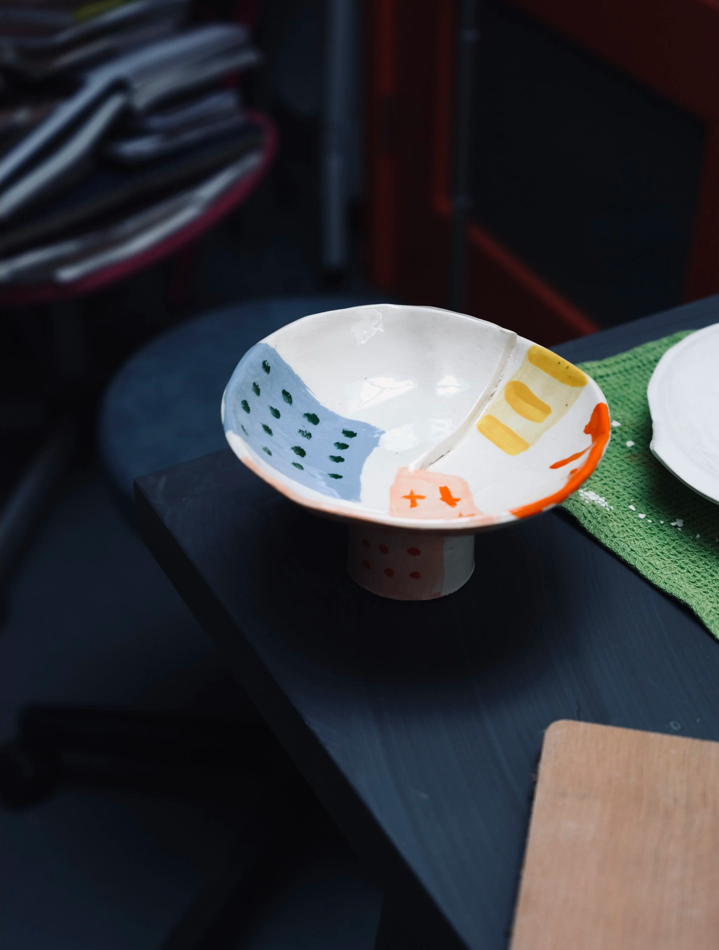 Close-up of a handmade painted ceramic bowl on a black surface with a green textured cloth underneath, in a room with a blurred background including stacked newspapers, a black speaker, and red furniture.