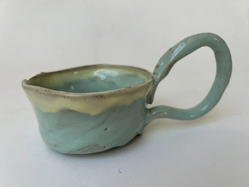 A small handmade ceramic cup with a curved handle, glazed in pastel shades of blue, green, and yellow.