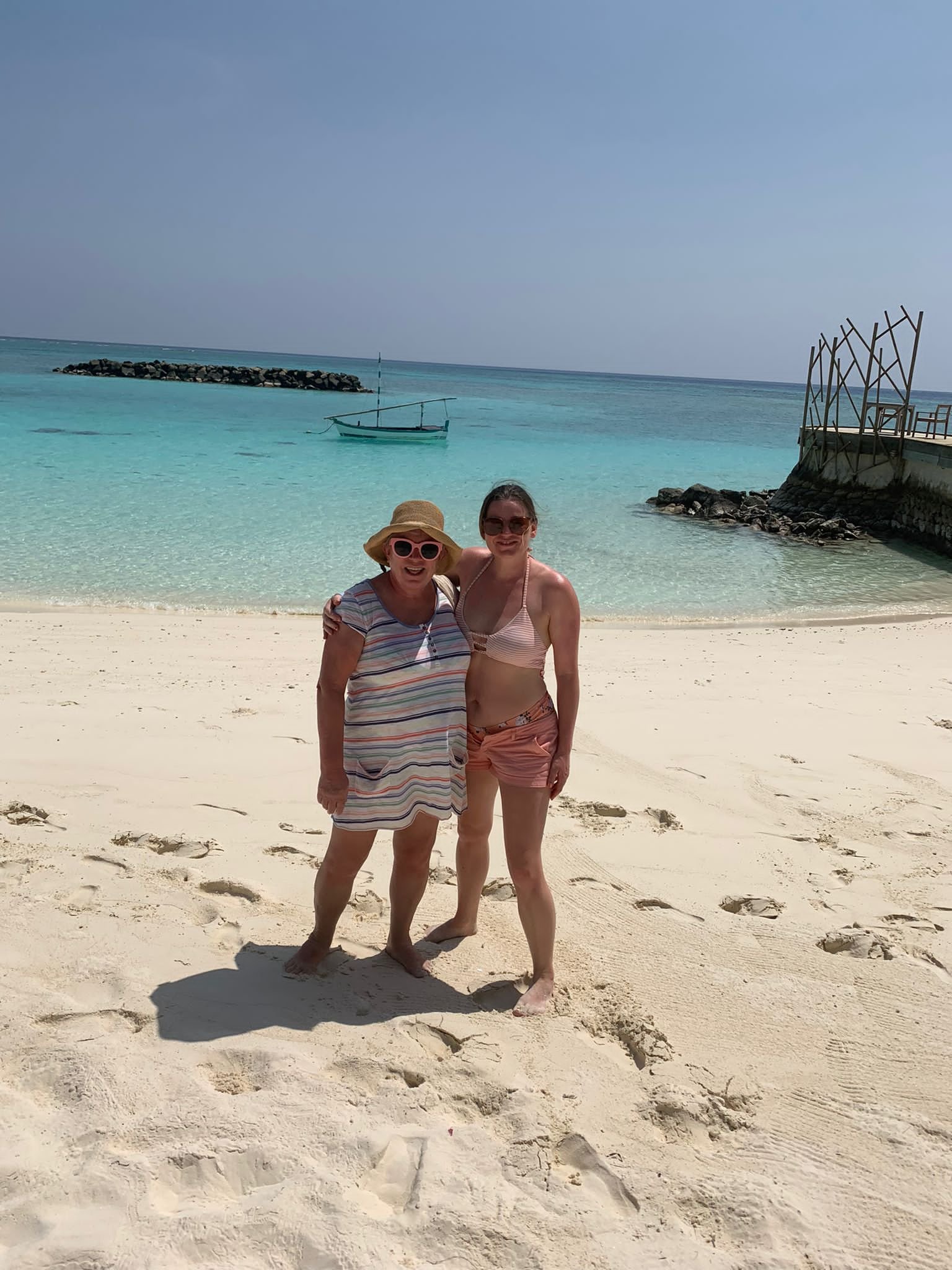 Two women standing barefoot on a sandy beach, smiling, with the turquoise ocean and a small boat in the background.