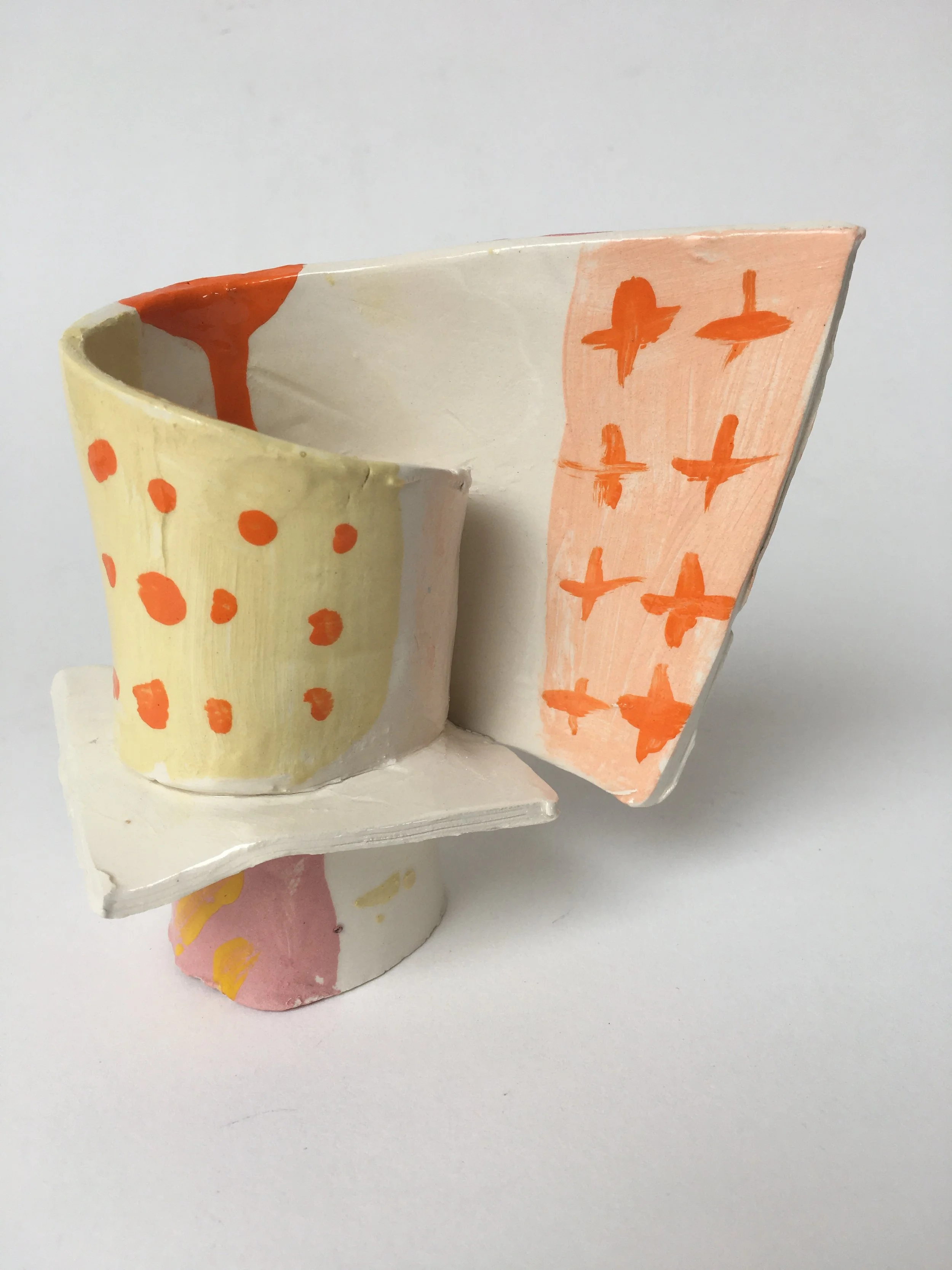 A small, abstract, multicolored sculpture with painted patterns of orange dots and bird-like shapes set against a white background.