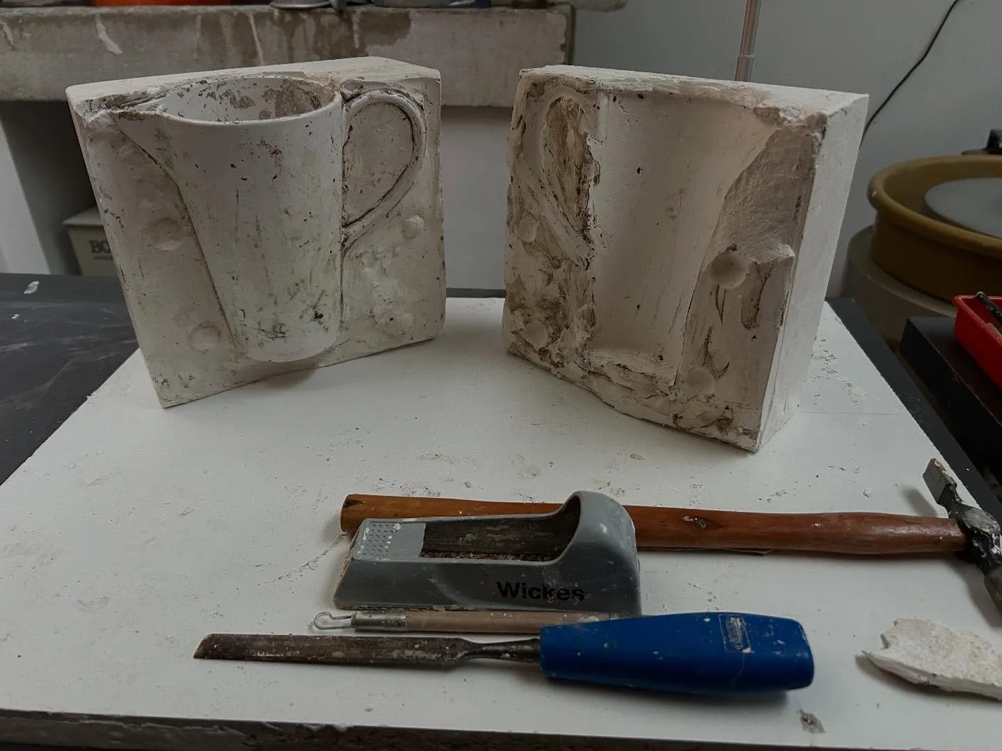 Two rectangular blocks of plaster or clay with carved relief designs, a hammer with the name ‘Wickes’ on it, a blue-handled tool, a chisel, and a small piece of broken material on a white work surface.