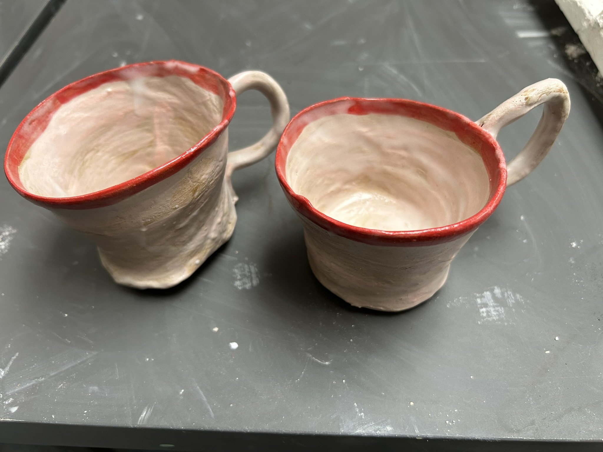 Two handmade ceramic mugs with cream-colored bodies and red rims, placed on a dark, slightly textured surface.