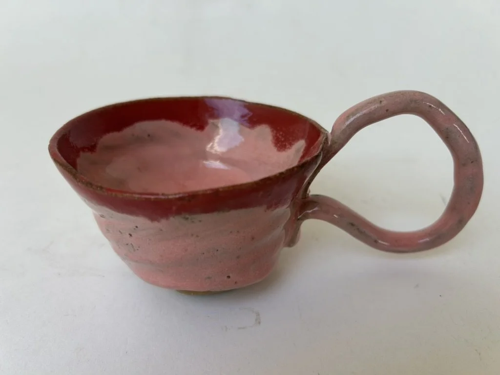 A small ceramic cup with a pink hue and a round handle, sitting on a white surface.