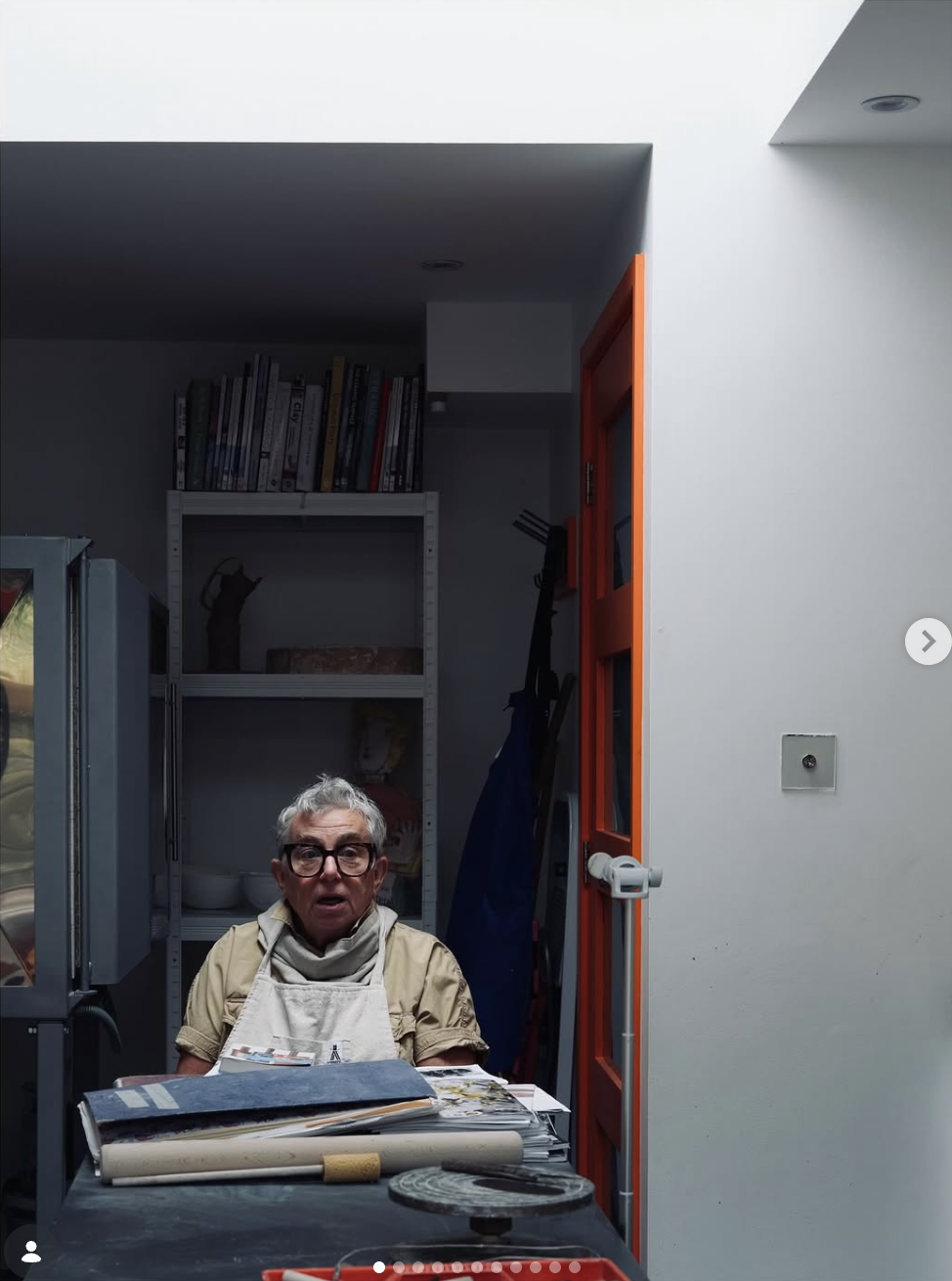 An elderly woman with short gray hair, glasses, and a beige shirt, sitting at a cluttered table in a room with white walls and a door with an orange frame. She appears surprised or confused, surrounded by various papers, a rolled-up poster, and objects on the table. Behind her is a shelf filled with books and decorative items.