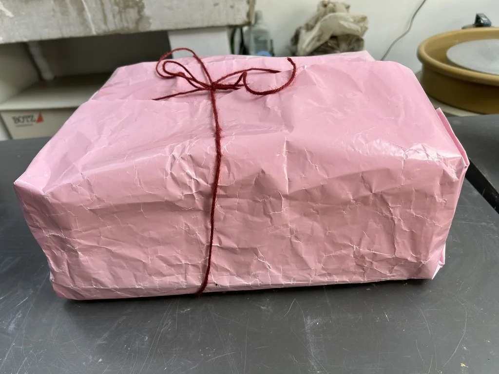 A rectangular object wrapped in wrinkled pink tissue paper, tied with a red string in a bow on top, placed on a black surface with a blurred background.
