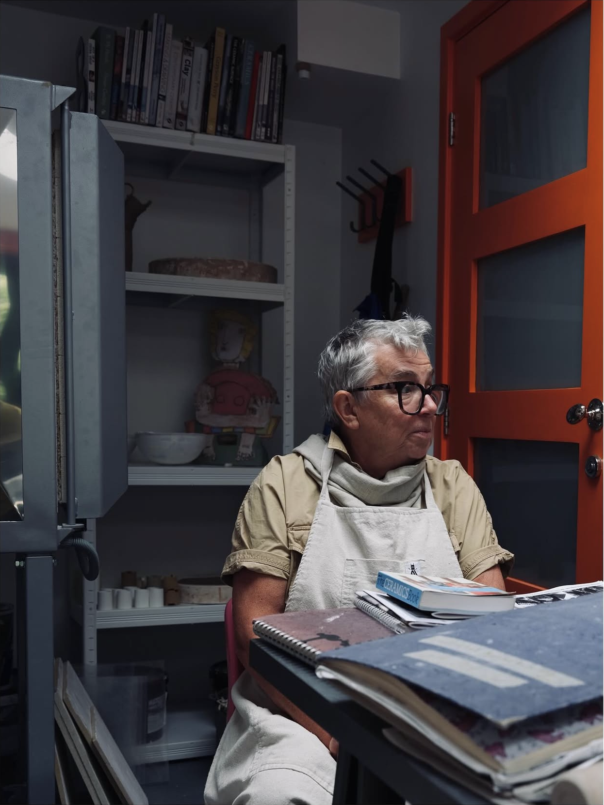 A woman with gray hair, glasses, and a beige shirt wearing an apron, sitting at a cluttered table in a room with shelves and an orange door in the background.