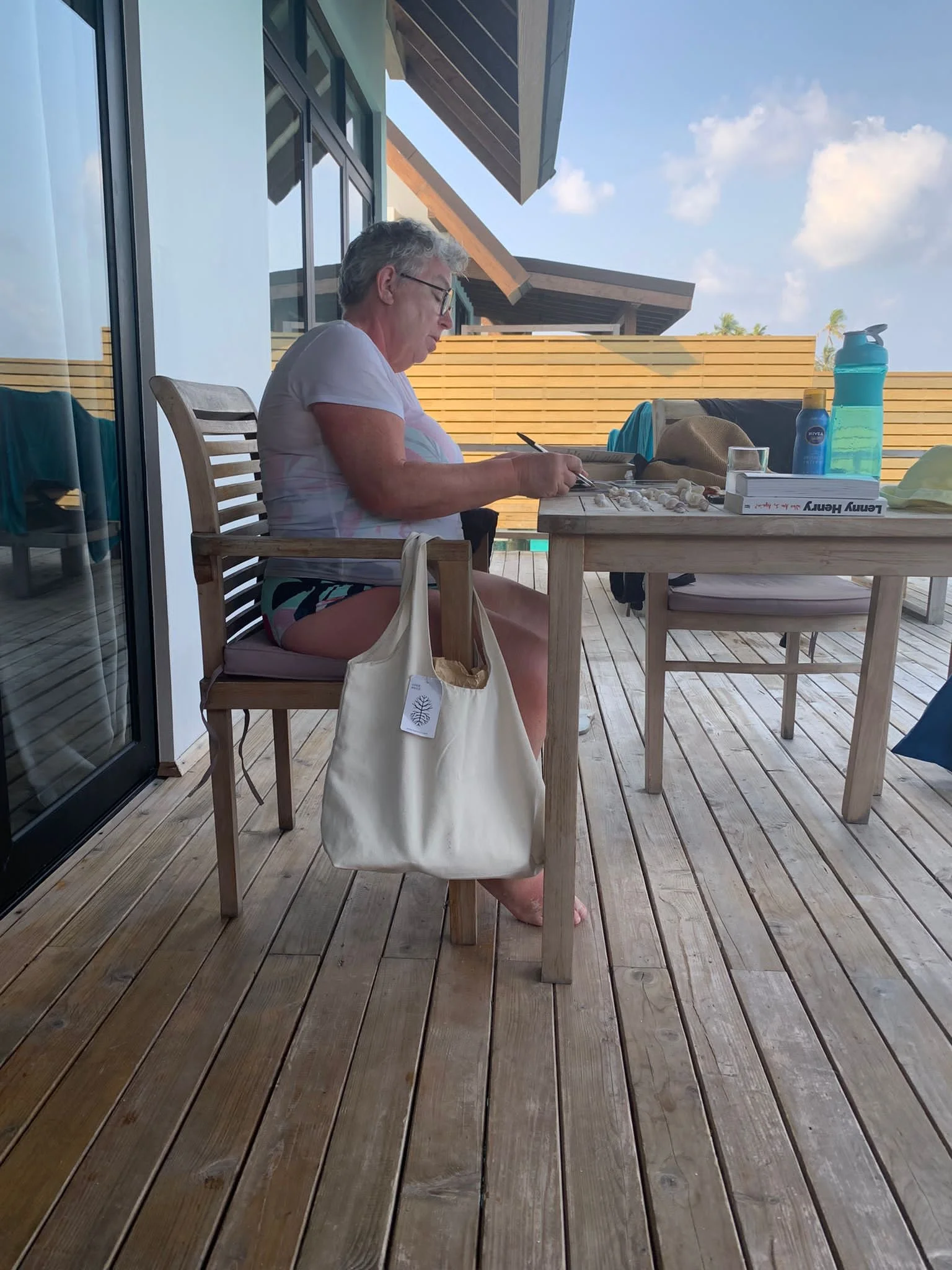 An older woman with gray hair and glasses sitting at a wooden outdoor table, writing or drawing, on a patio with a wooden fence and blue sky in the background.