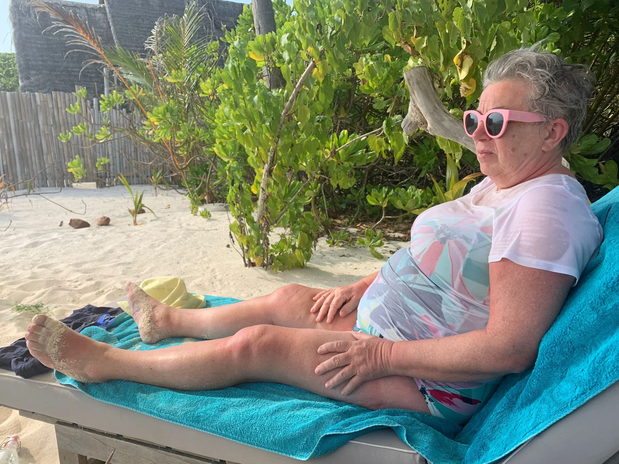 An elderly woman with short gray hair wearing pink sunglasses and a pastel-colored T-shirt relaxing on a beach chair under greenery with sand and a wooden fence in the background.