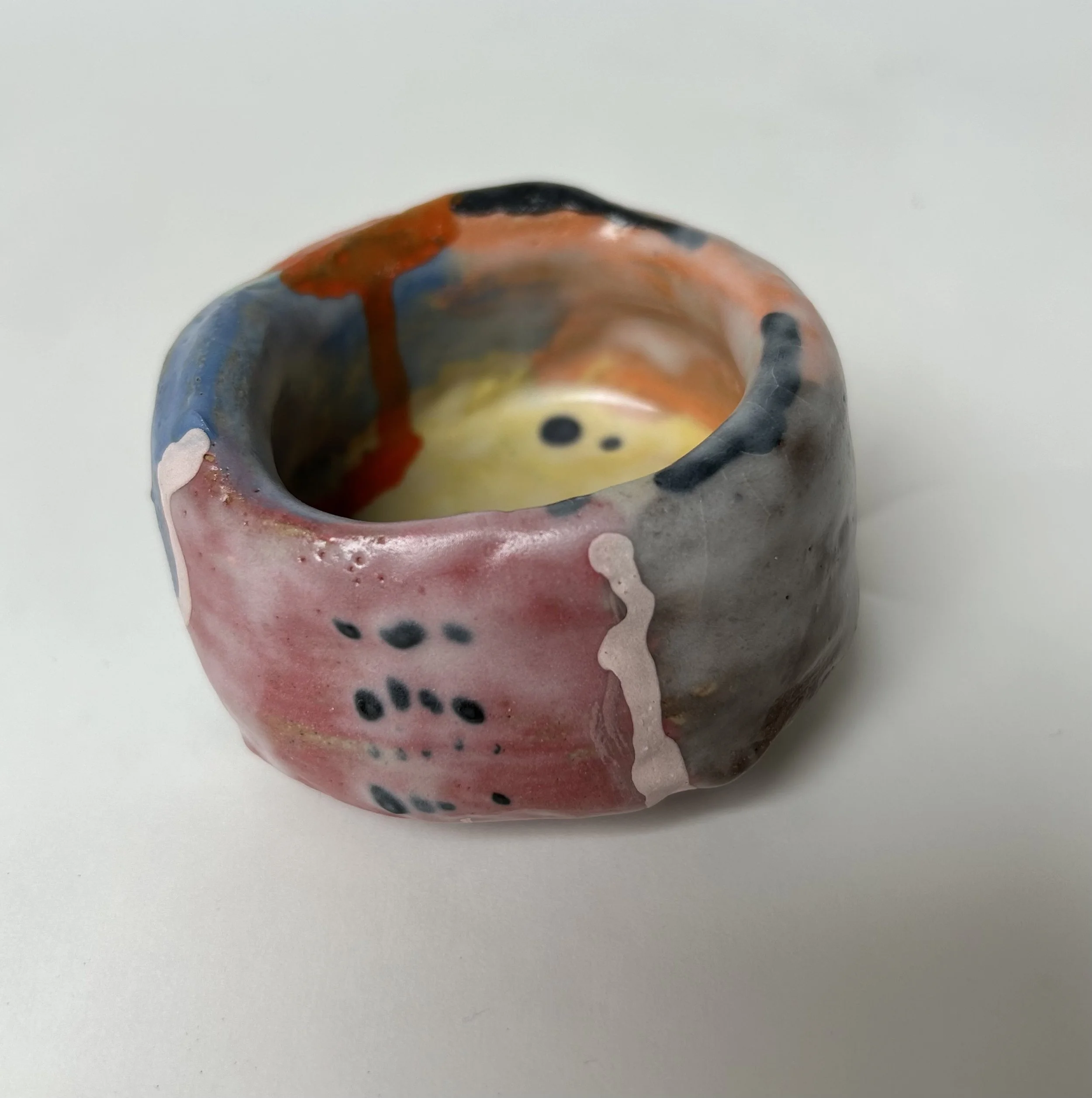 Close-up of a small, colorful, handcrafted ceramic or clay art piece with a hole on top, featuring a multicolored glaze with black, red, pink, and blue hues, placed on a plain white surface.