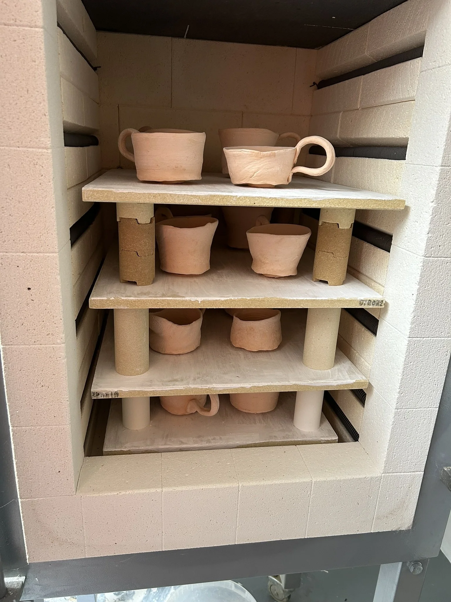 Unfinished clay cups and bowls on shelves in a kiln or oven, with ceramic supports and beige bricks surrounding.