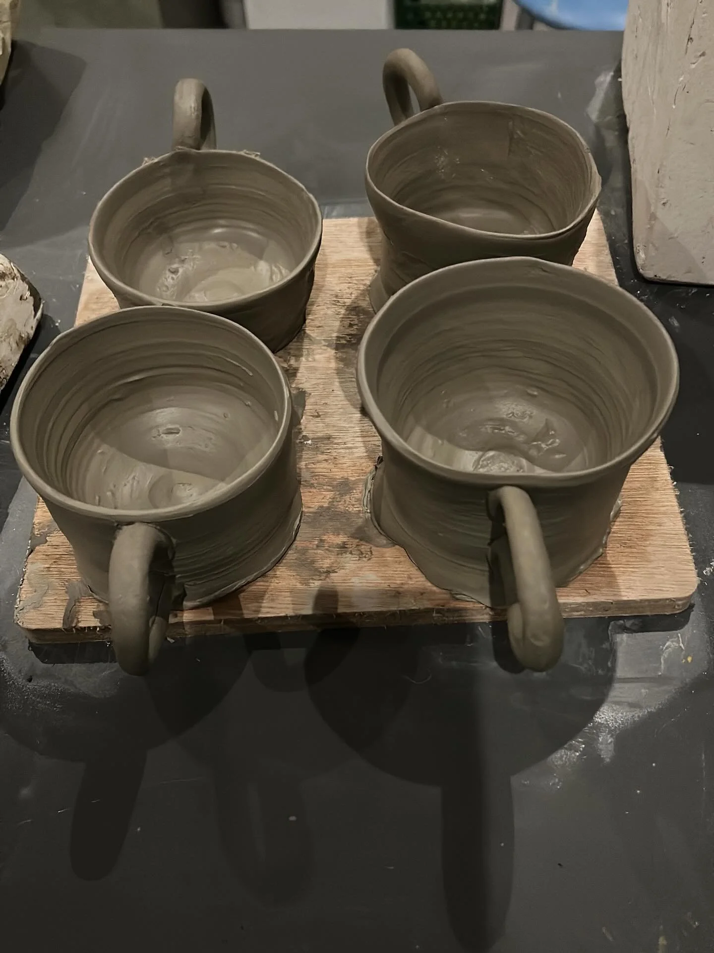 Four unfinished clay coffee mugs with handles on a wooden board in a pottery studio.
