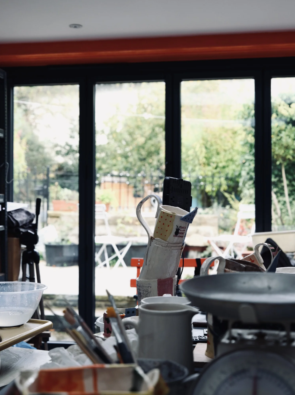 Indoor scene with cluttered table, baking tools, mugs, and bowls, with large windows outside showing a garden or backyard with trees and patio furniture.