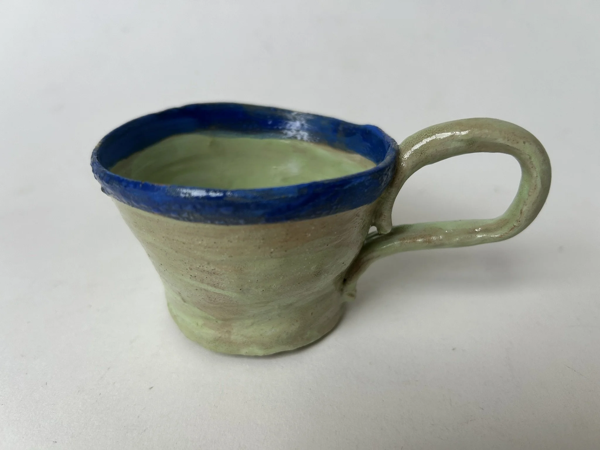 A ceramic cup with a handle, featuring a blue rim and an interior that appears to have a greenish glaze. The cup has a textured surface with natural color variations.