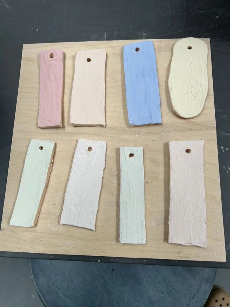 Colored wooden paint samples on a light wooden board, arranged in two rows, with pastel colors including pink, beige, blue, yellow, green, white, and pale pink.