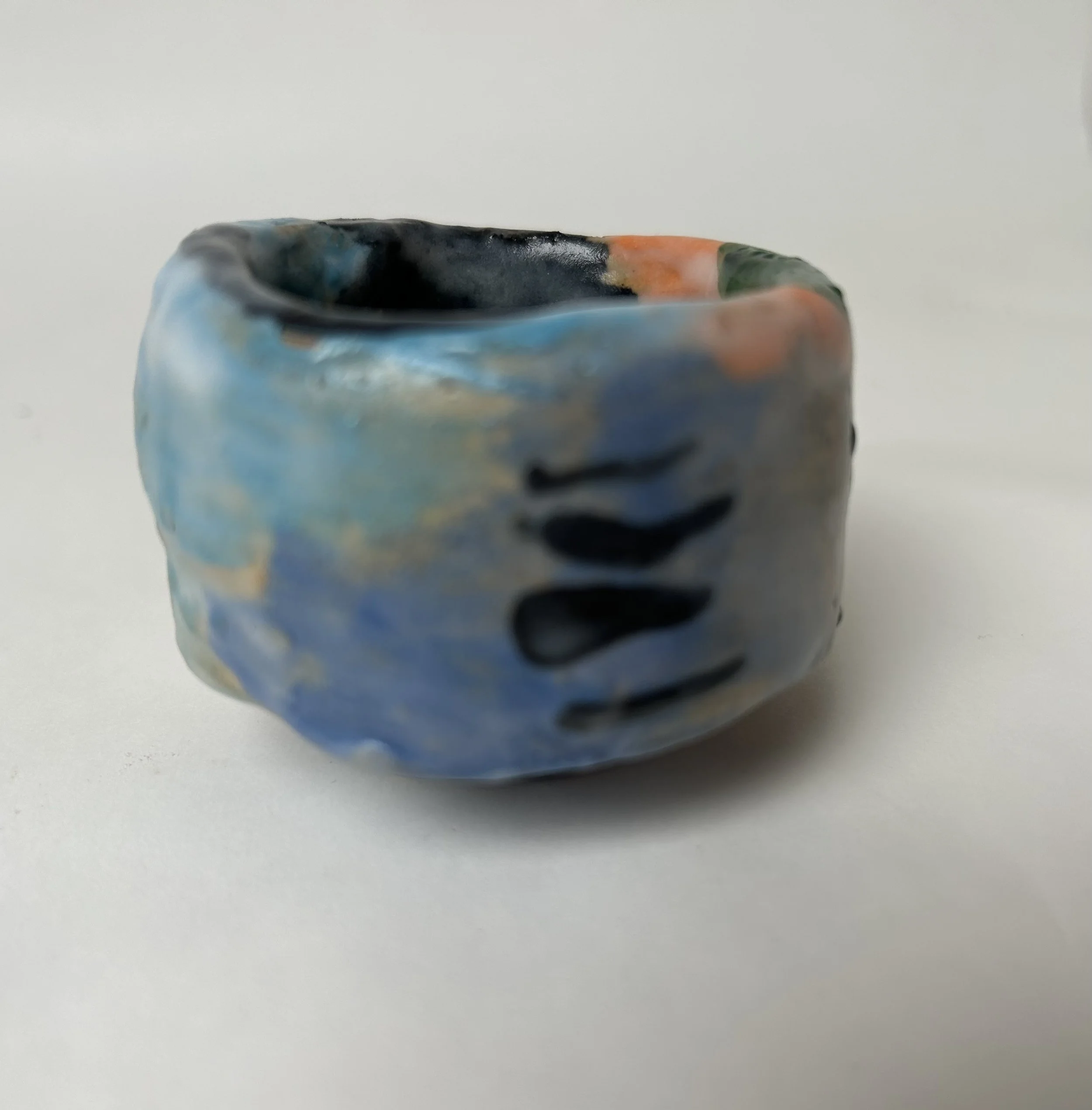 Close-up of a small ceramic bowl with a marbled blue, black, orange, and green glaze, set against a plain gray background.