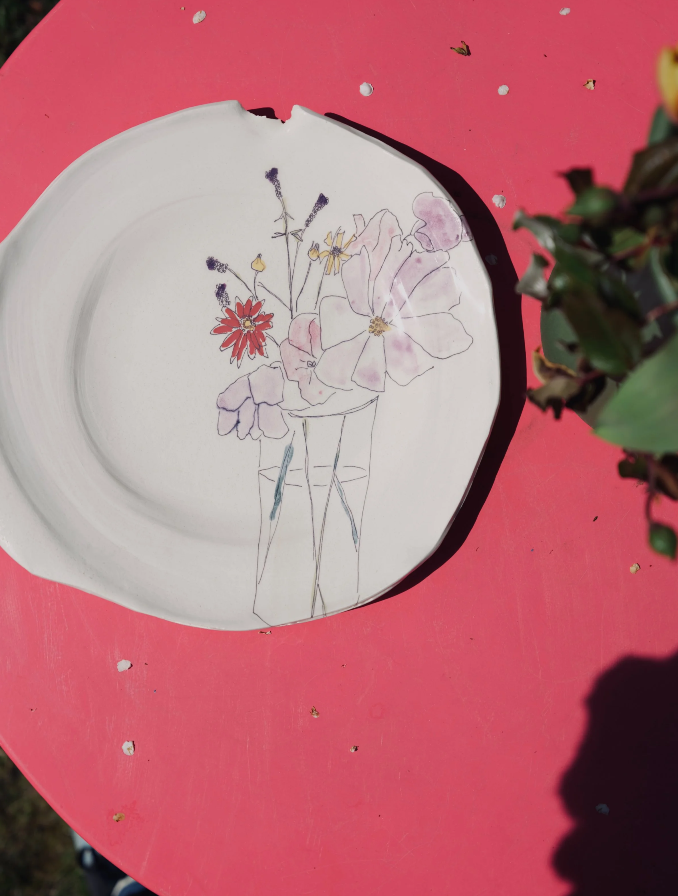 A white oval ceramic plate with a floral illustration in single lines and delicate watercolor accents, featuring large flowers and small buds. The plate is placed on a pink surface with some debris and a blurred plant to the right. The plate has a small chip on the upper edge.