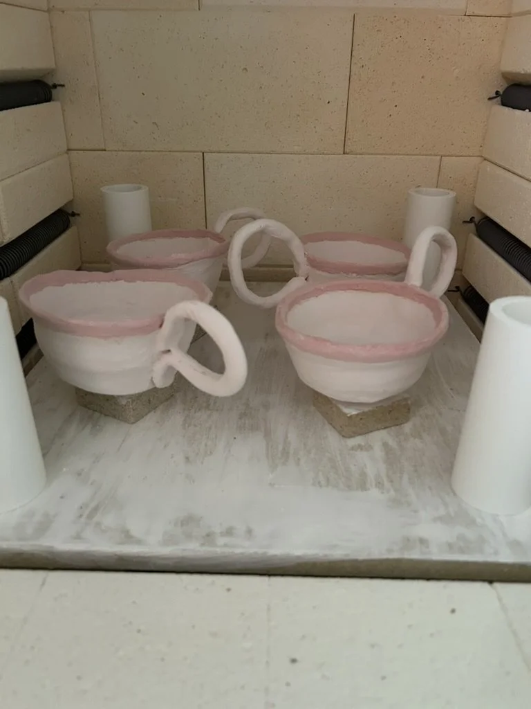 Decorative ceramic cups with handles, placed on small pedestals on a white surface, resembling cups of tea or coffee, with a pink rim detail, set against a beige tiled wall.