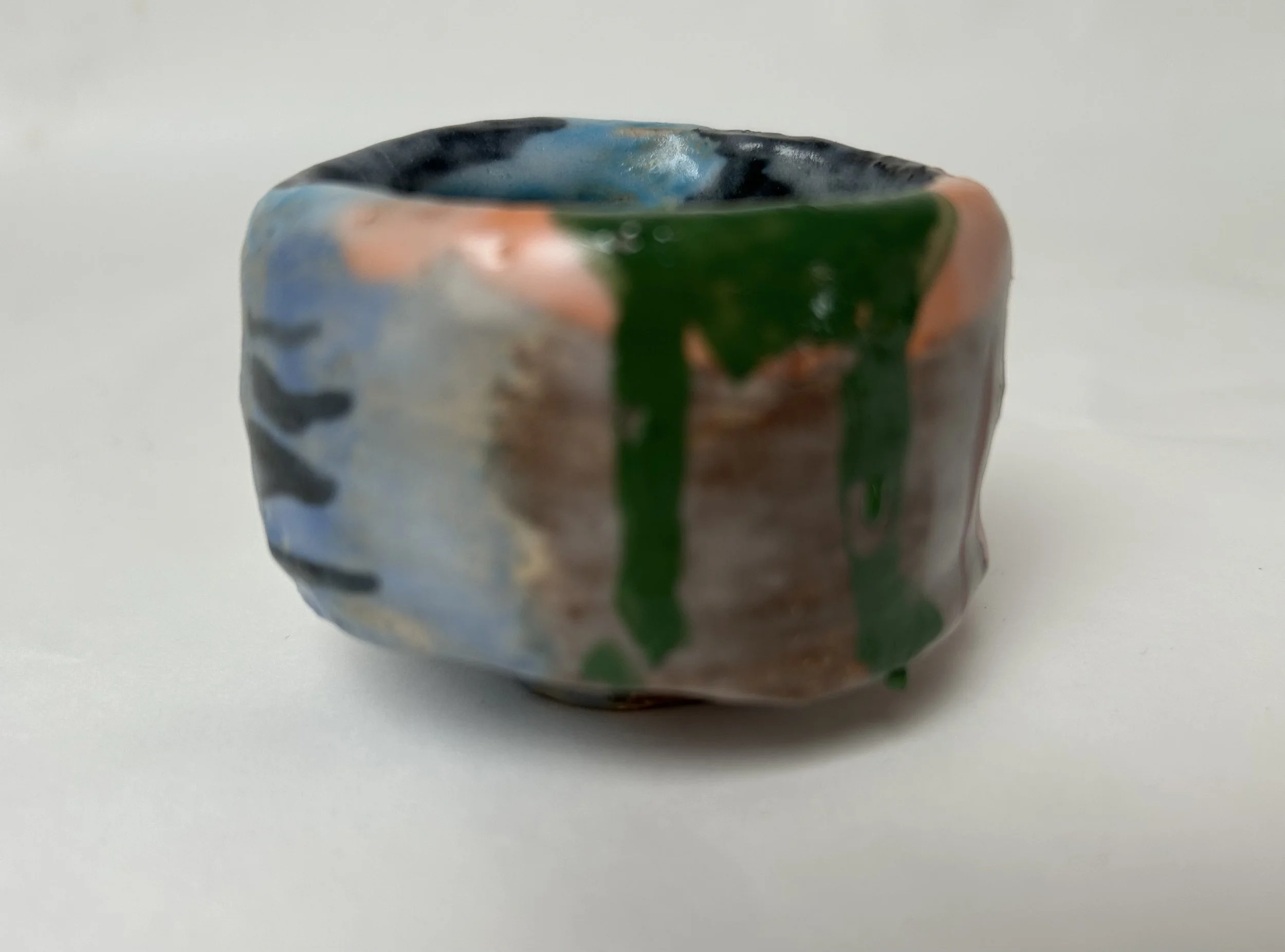 Close-up of a small, multicolored ceramic cup with a glossy finish, featuring black, blue, green, and peach tones, positioned on a plain white surface.
