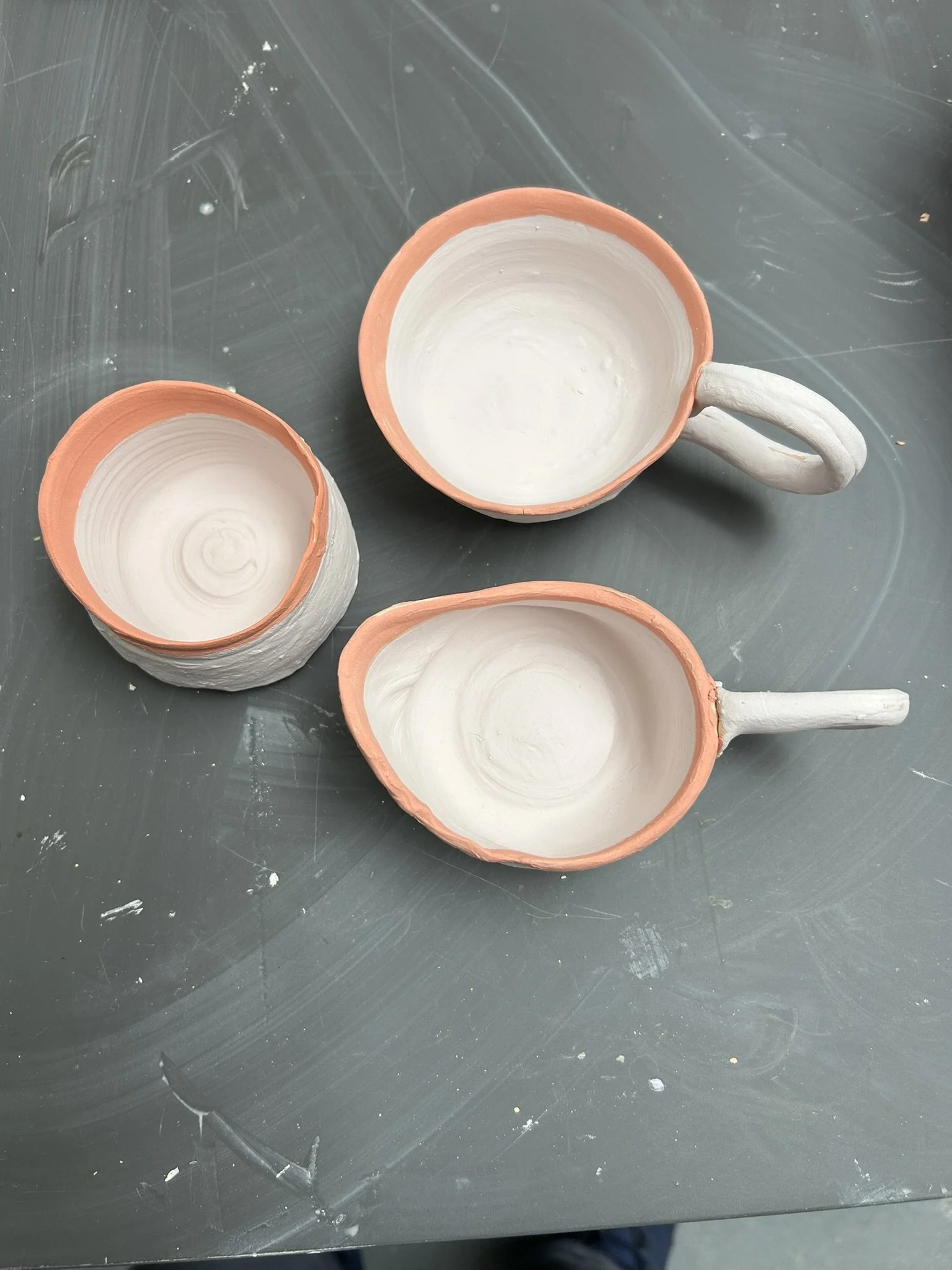 Three unglazed ceramic cups with handles on a gray surface with visible chalk dust and marks.