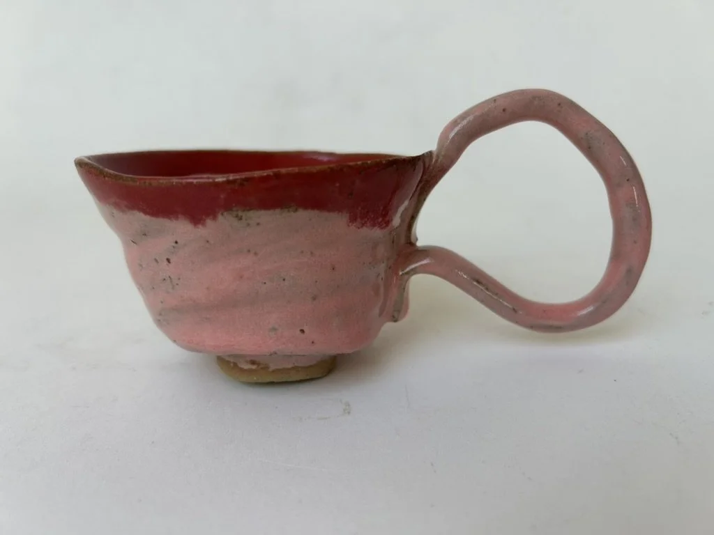 A small ceramic cup with a pink glaze and a round handle, placed on a white surface.