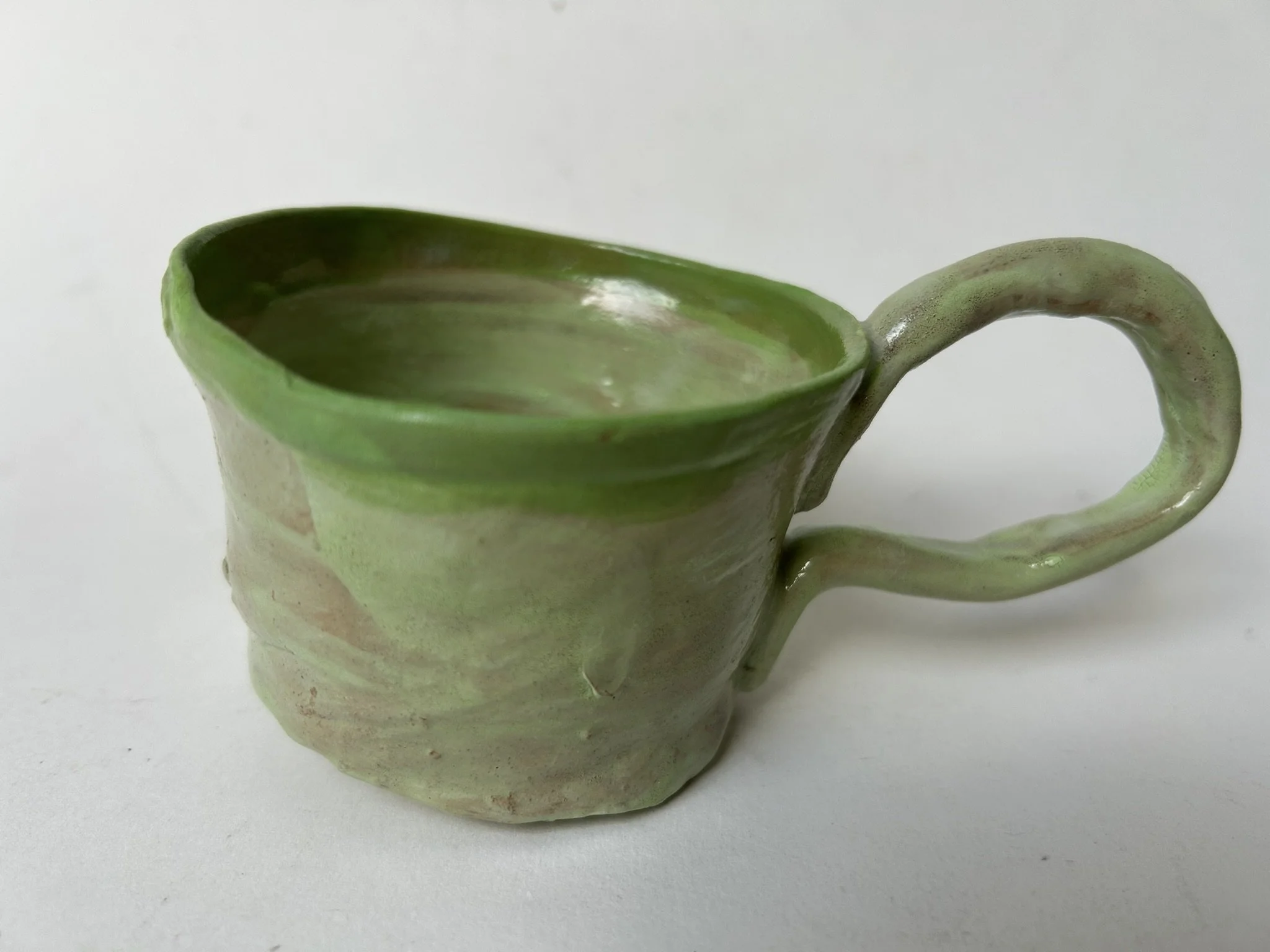 Close-up of a ceramic mug with a green interior and a curved handle, featuring a pastel green and pink glaze finish, against a plain white background.