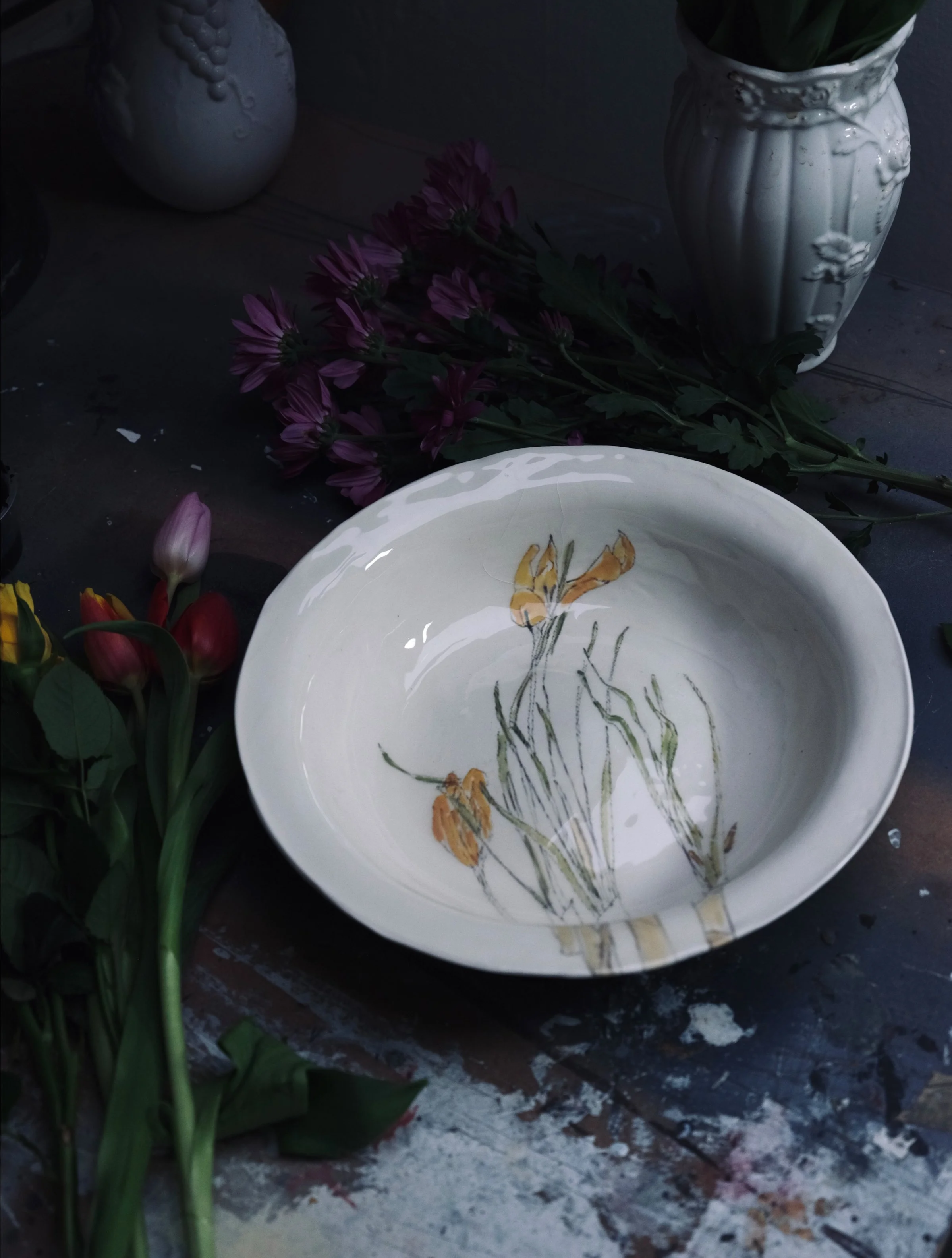 A still life arrangement with a white ceramic vase filled with green plant leaves, a white decorative vase, a white ceramic plate with a painted flower, purple flowers, and some tulips on a dark wooden surface.