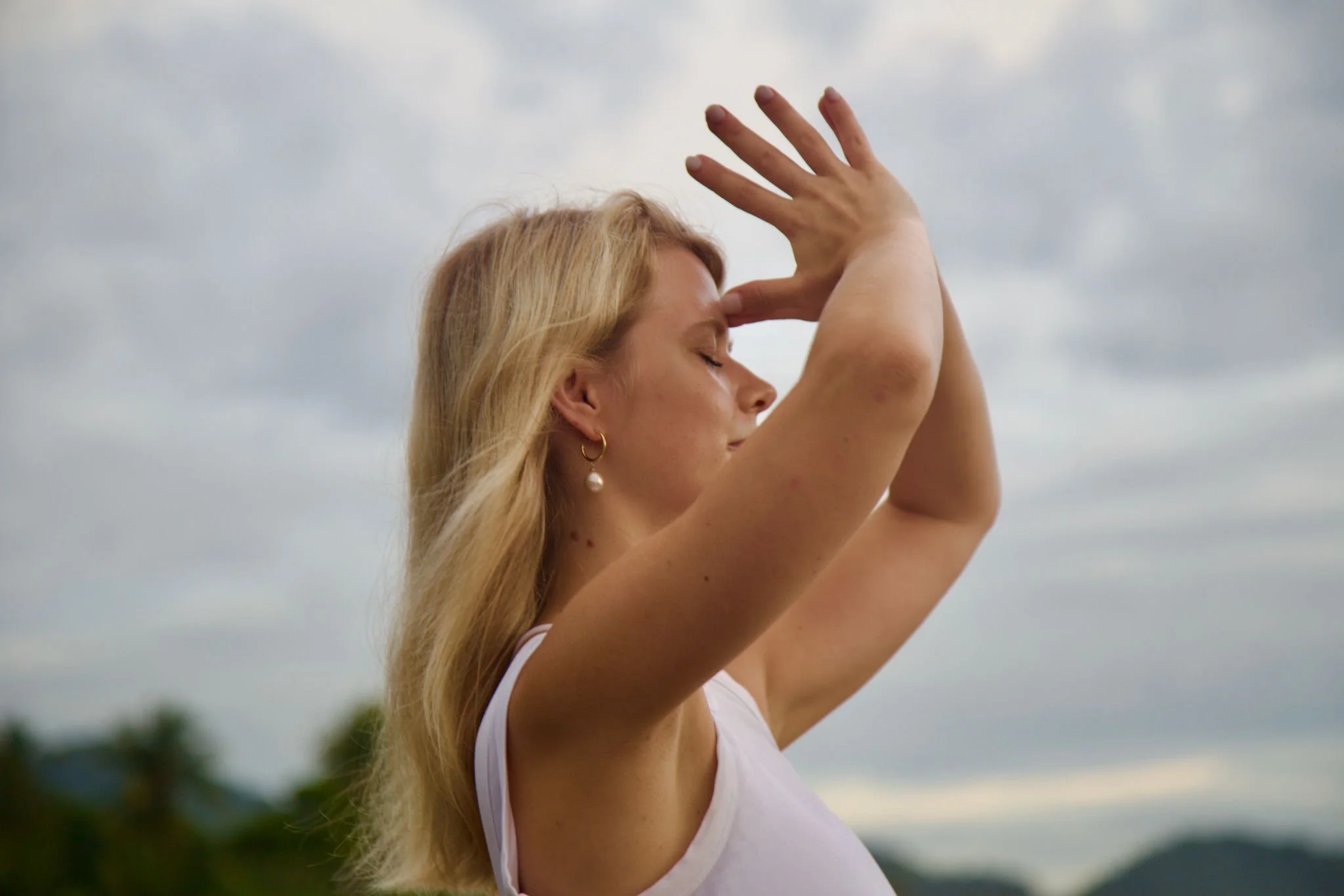 Eine blonde Frau mit geschlossenen Augen und einem weißen Top macht eine Yoga- oder Meditationsübung im Freien bei bewölktem Himmel.