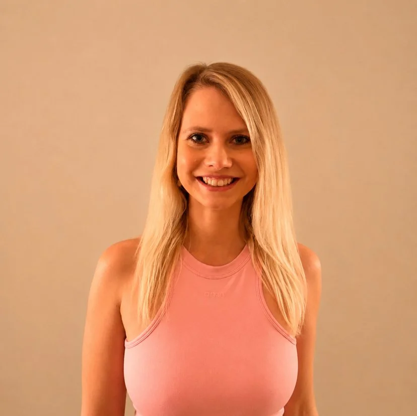 Eine lächelnde blonde Frau in einem pinken ärmellosen Top vor einem neutralen Hintergrund.