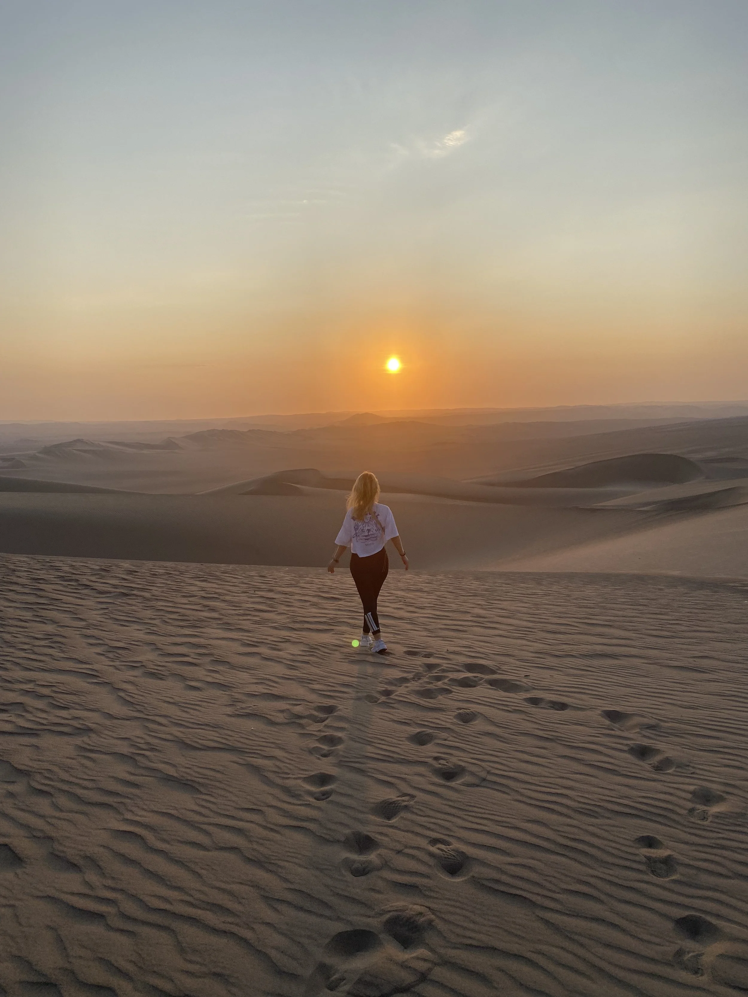 Eine Frau läuft in den Sanddünen bei Sonnenuntergang, der Himmel ist leicht bewölkt und die Landschaft ist weitgehend sandig.