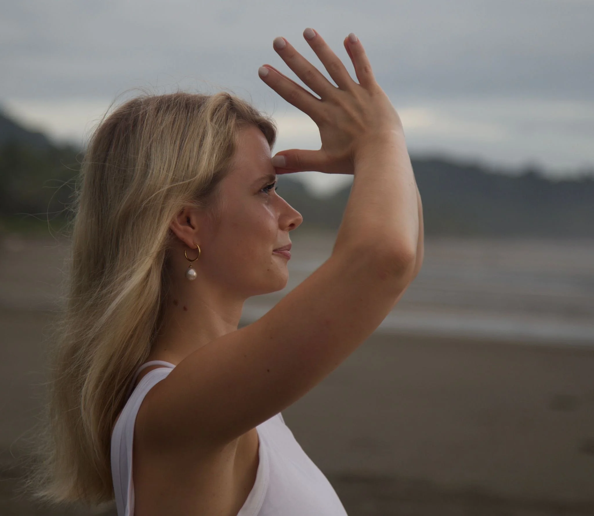 Eine Frau mit blonden Haaren und Ohrringen macht eine Yoga-Pose am Strand bei bewölktem Himmel.