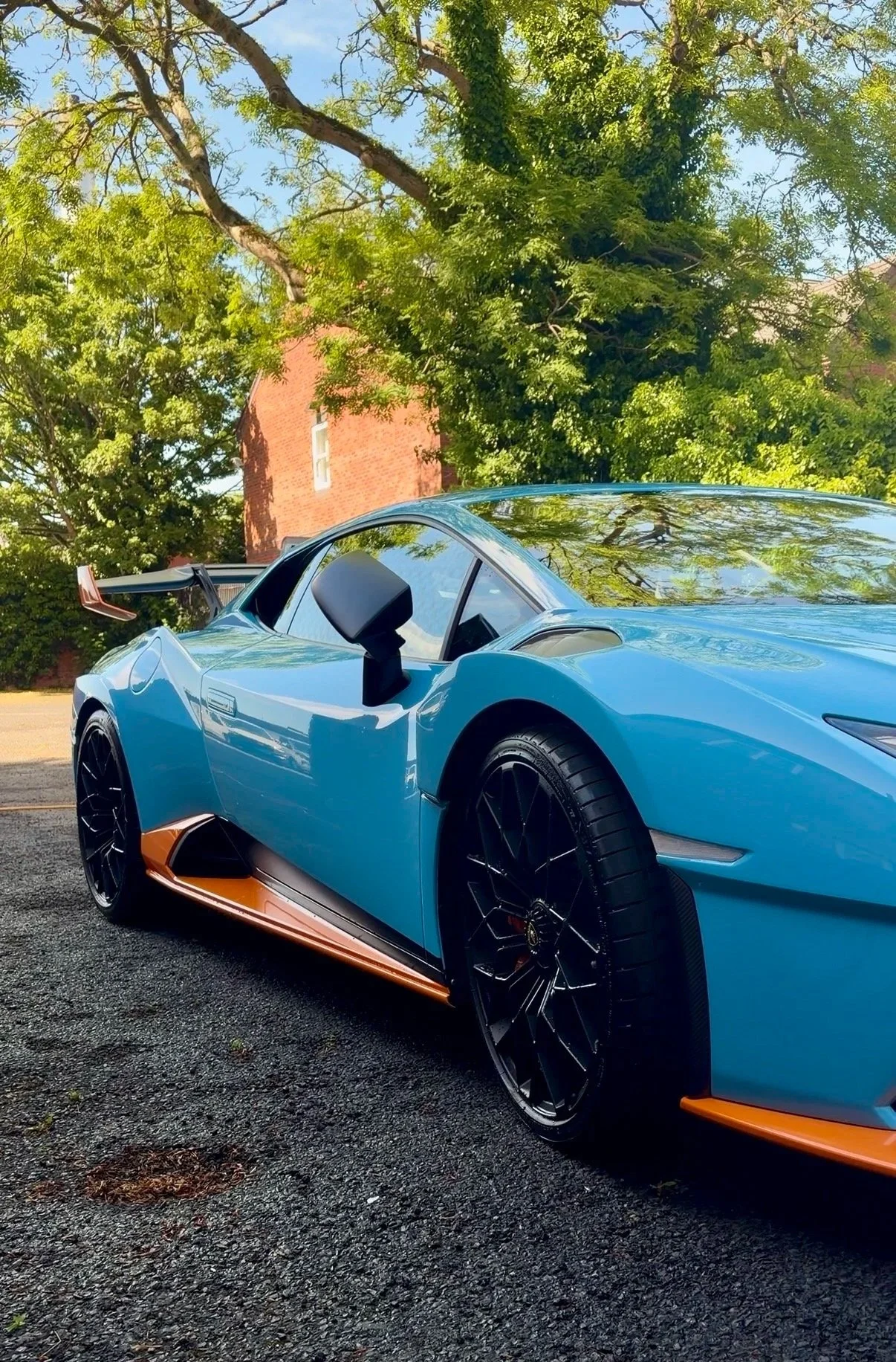 A blue sports car with black wheels and orange accents parked on a gravel surface under trees with green foliage and a brick building in the background.