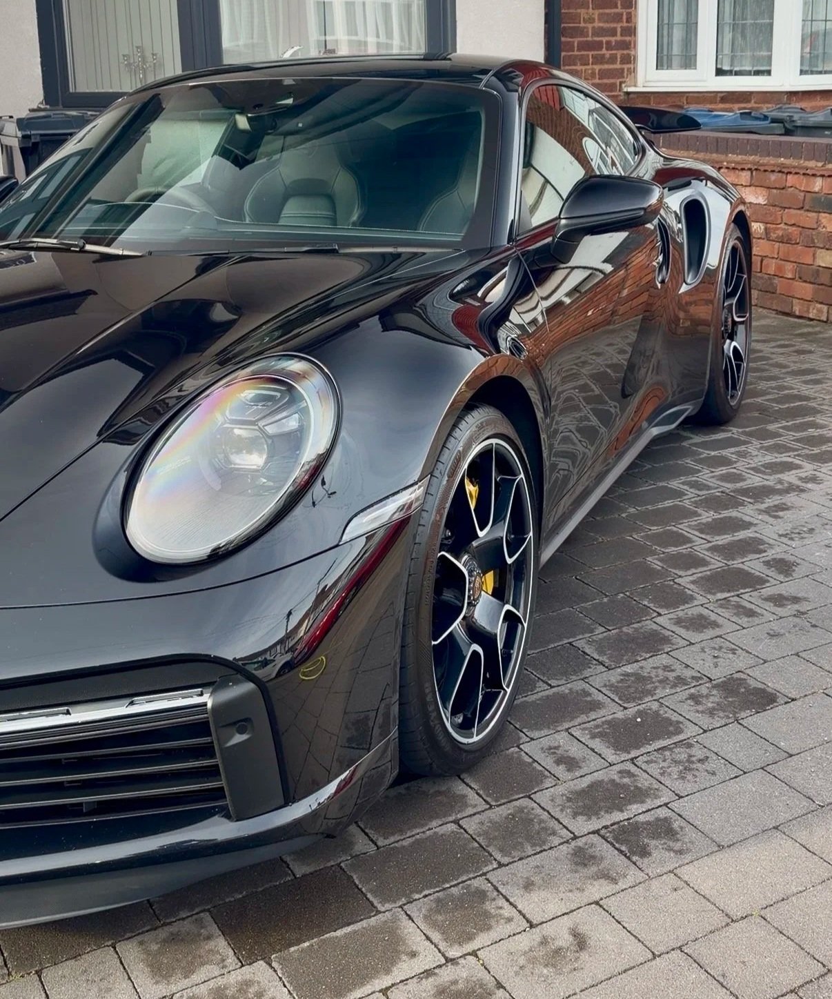 Black sports car parked on a brick driveway in front of residential houses with bay windows and brick walls.