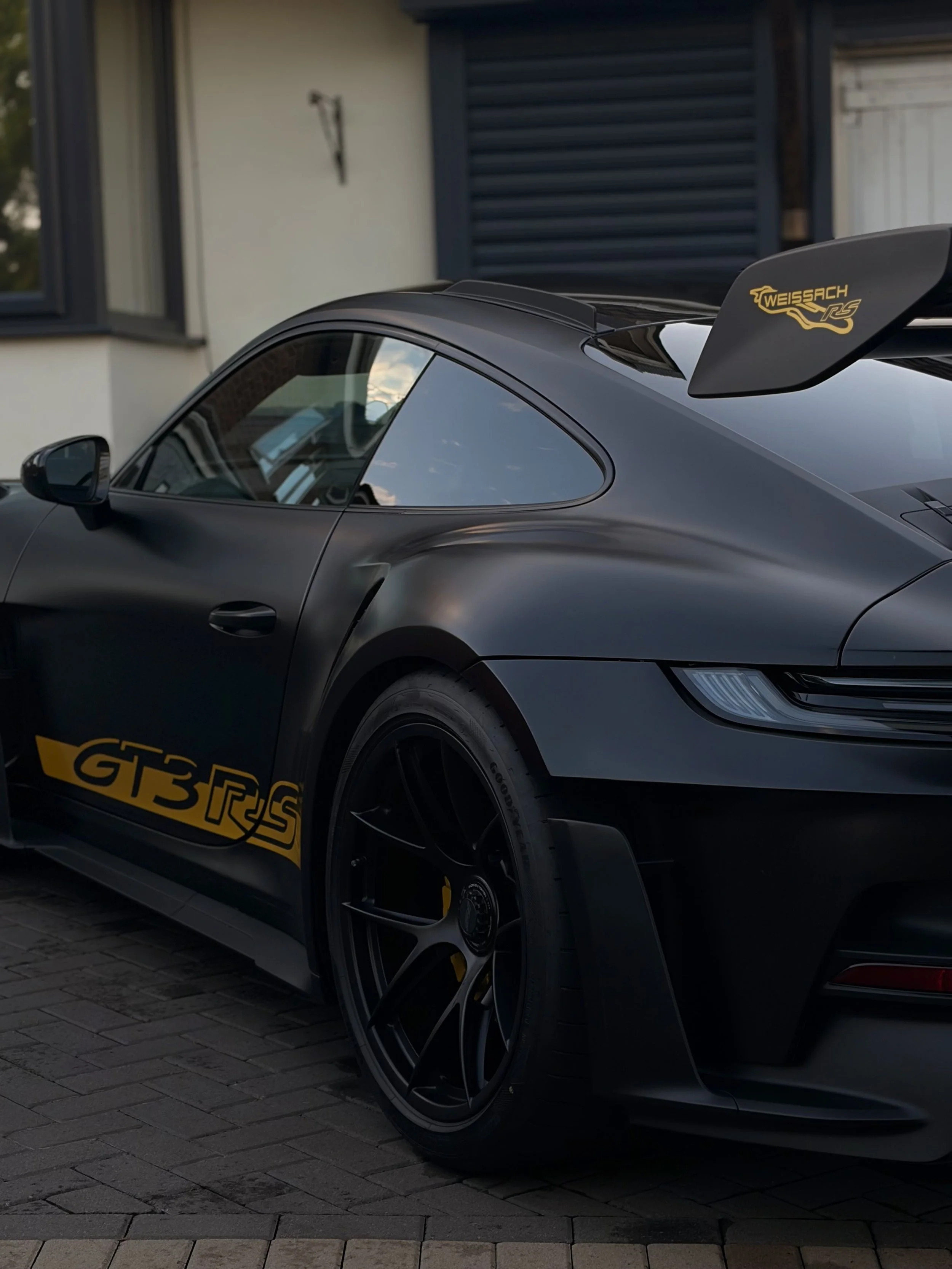 Black sports car with a yellow and black decal reading 'GT3 RS' on the side, parked on a driveway with houses in the background.