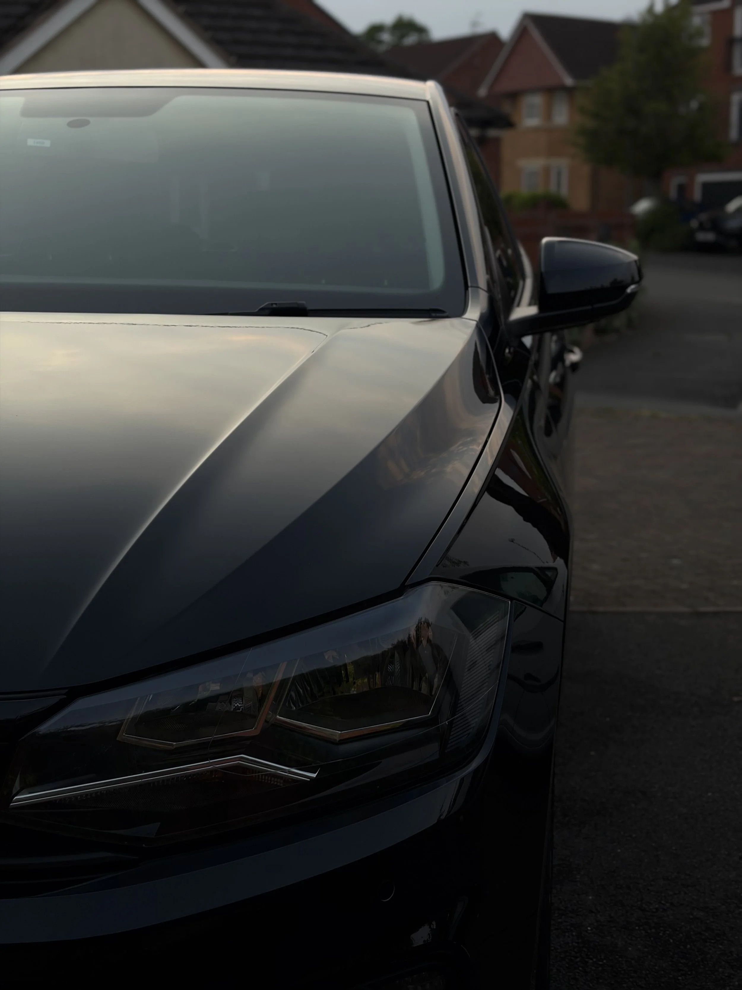 Close-up of the front part of a black car parked on the street in front of residential houses, with a focus on the windshield, headlight, and side mirror.