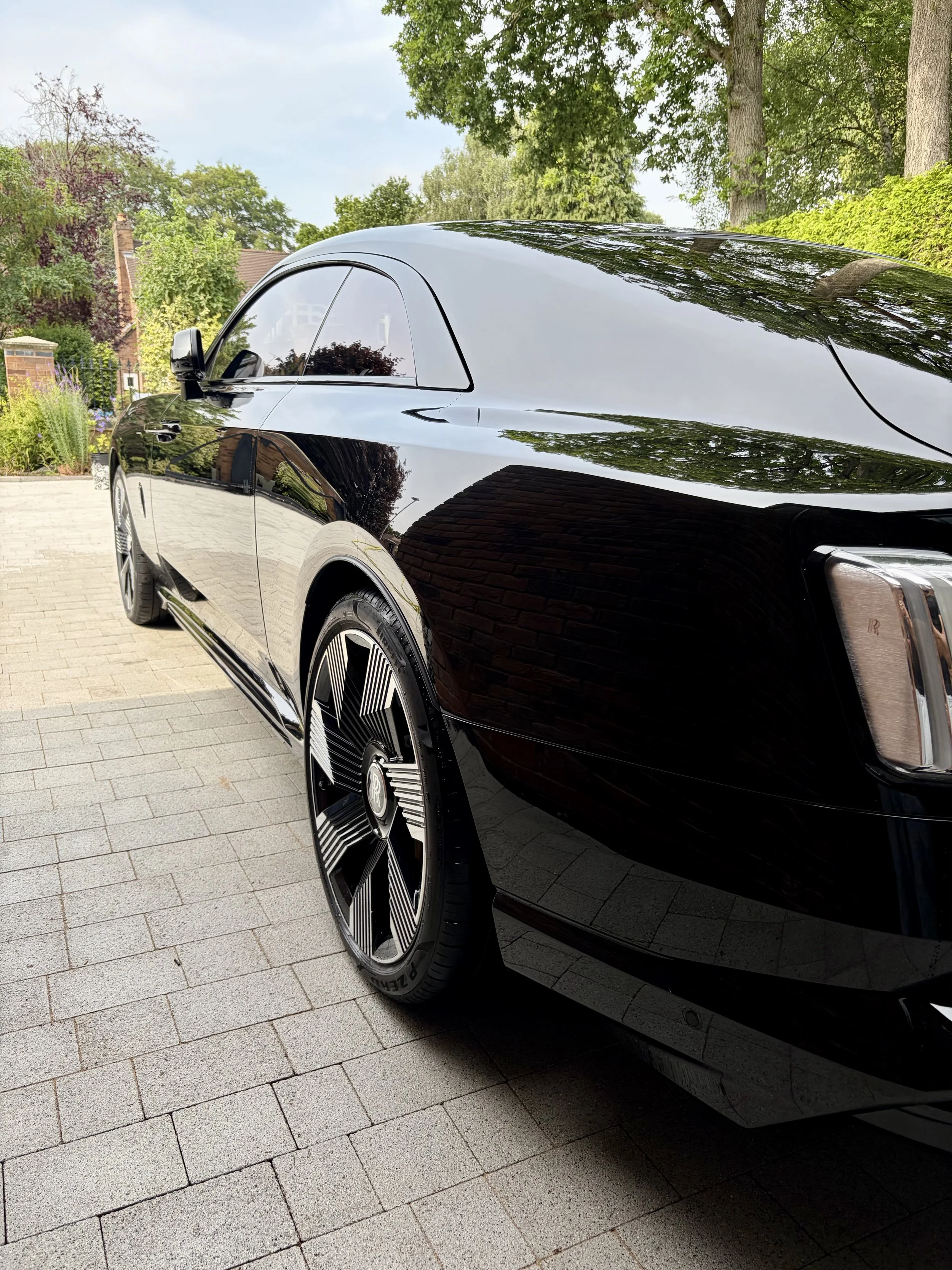 A sleek black luxury car parked on a paved driveway with green trees and a garden in the background.