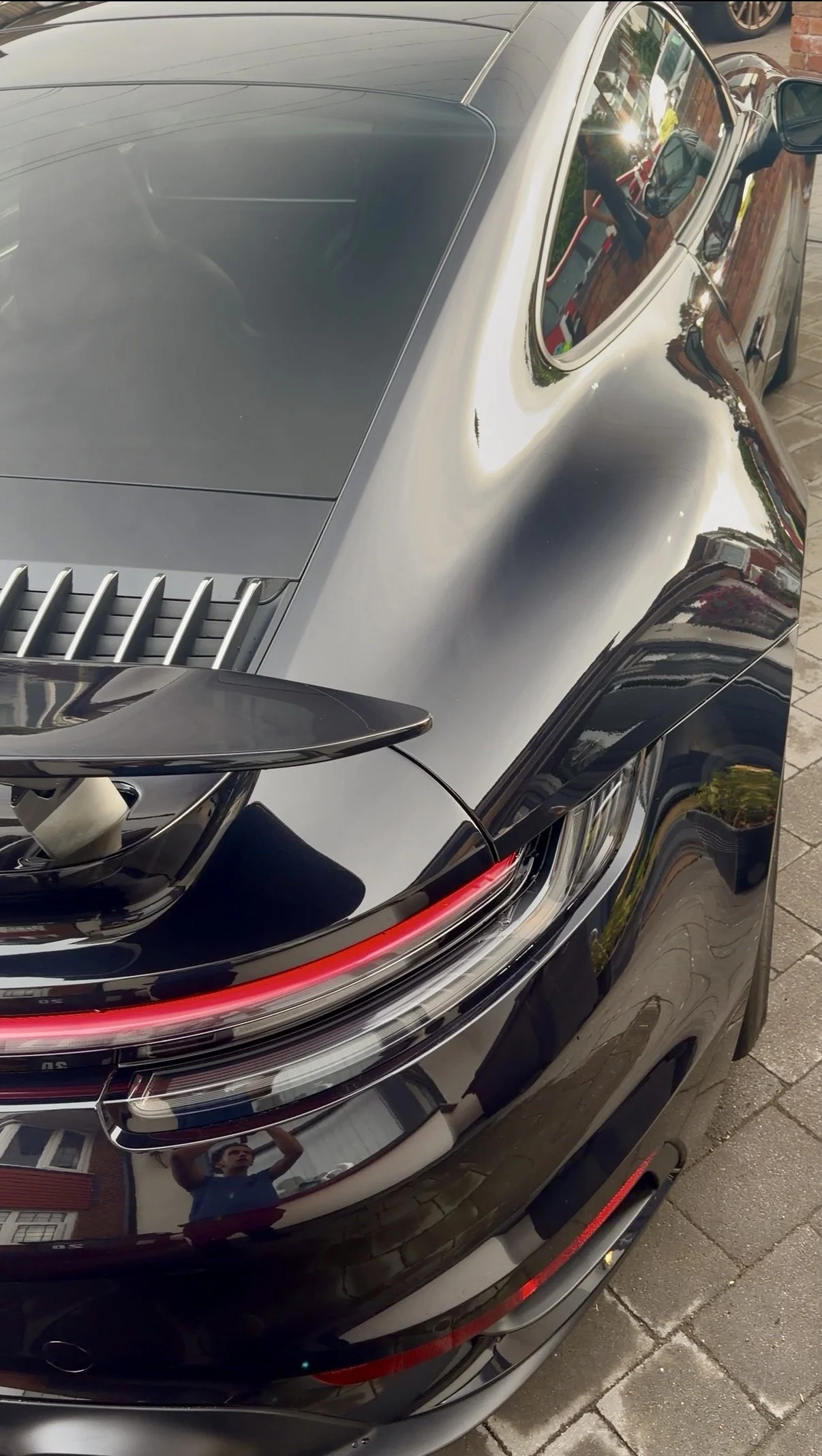 Close-up of the rear end of a black sports car with a curved spoiler, reflective surface, and taillight. The reflection shows a person taking the photo and nearby houses and sky.