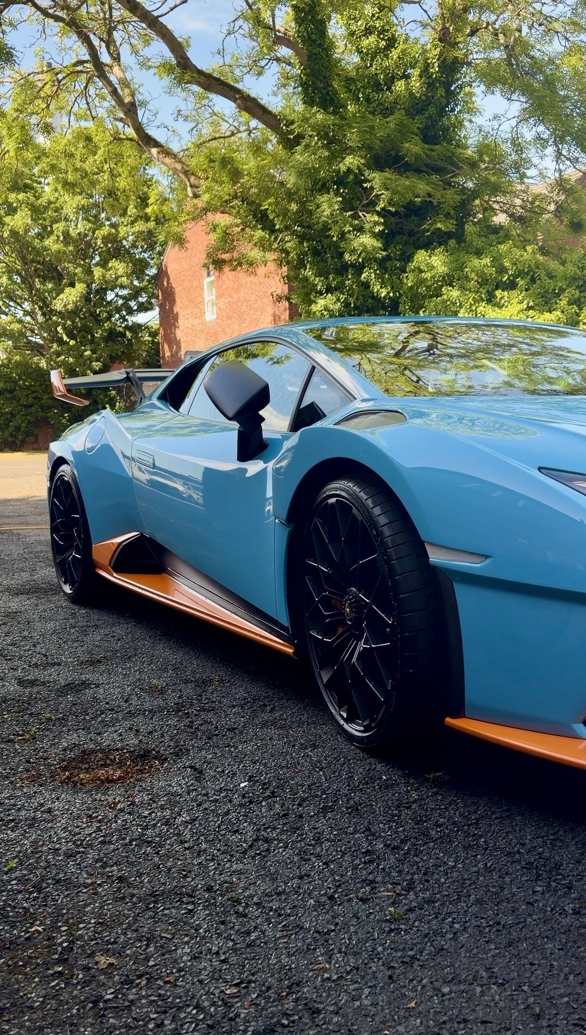 A blue sports car parked outdoors on a gravel surface, surrounded by green trees and a red brick building in the background.