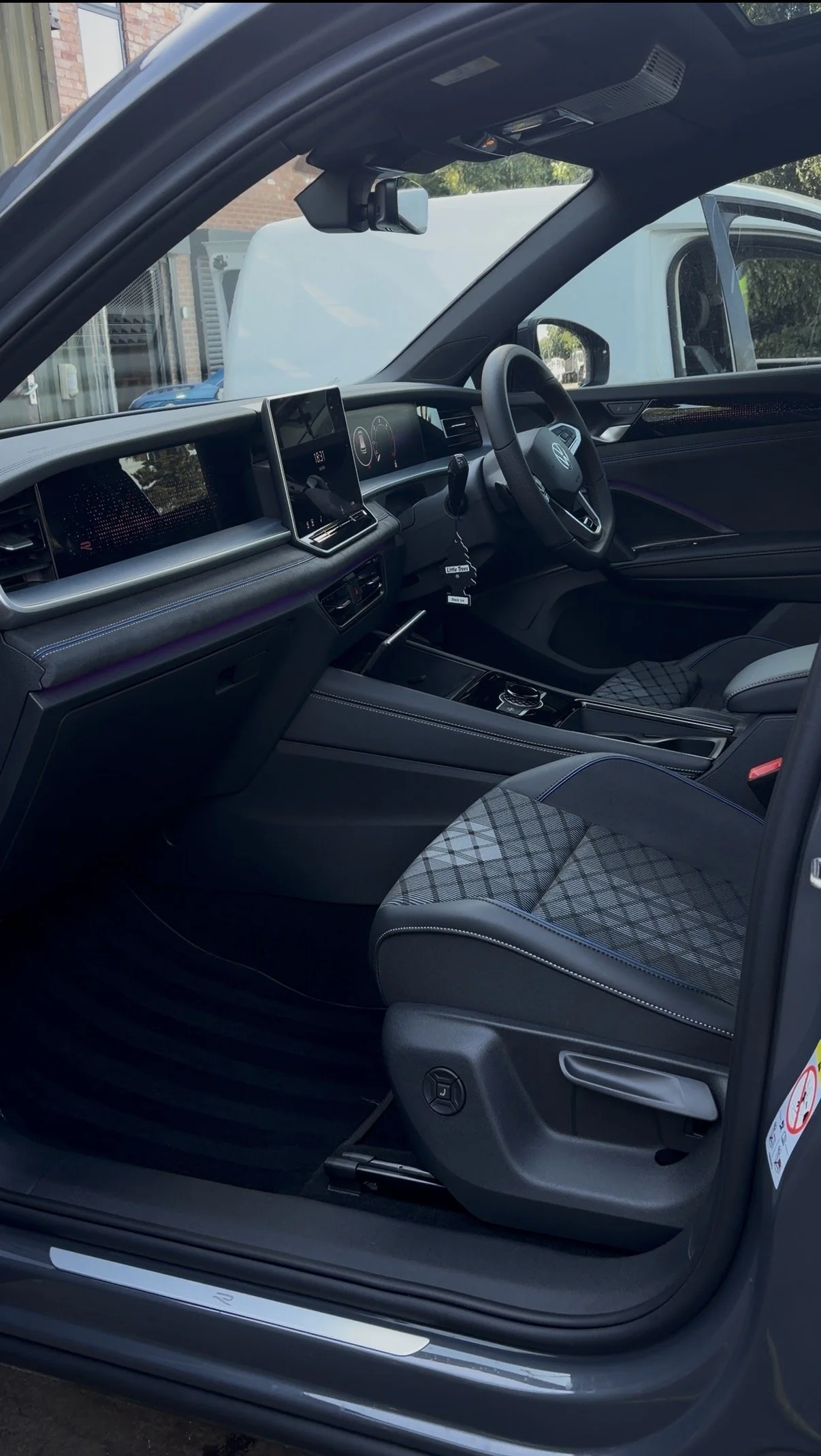 Interior of a modern Volkswagen car with a digital dashboard, a steering wheel, and a plaid fabric seat. The car appears to be parked outdoors.