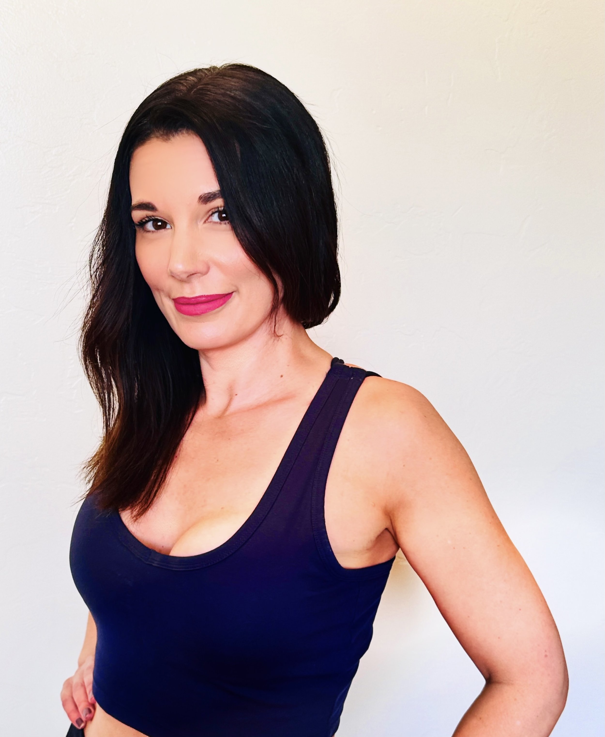 A woman with long dark hair and pink lipstick is smiling, wearing a black sleeveless top, standing against a plain white wall.
