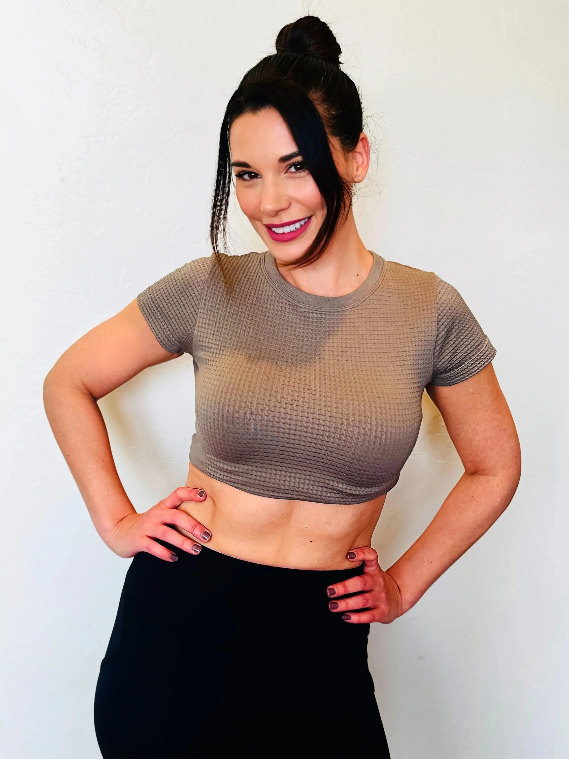 A woman in a beige crop top and black pants with dark hair styled in a top knot, smiling and resting her hands on her hips against a plain white wall.