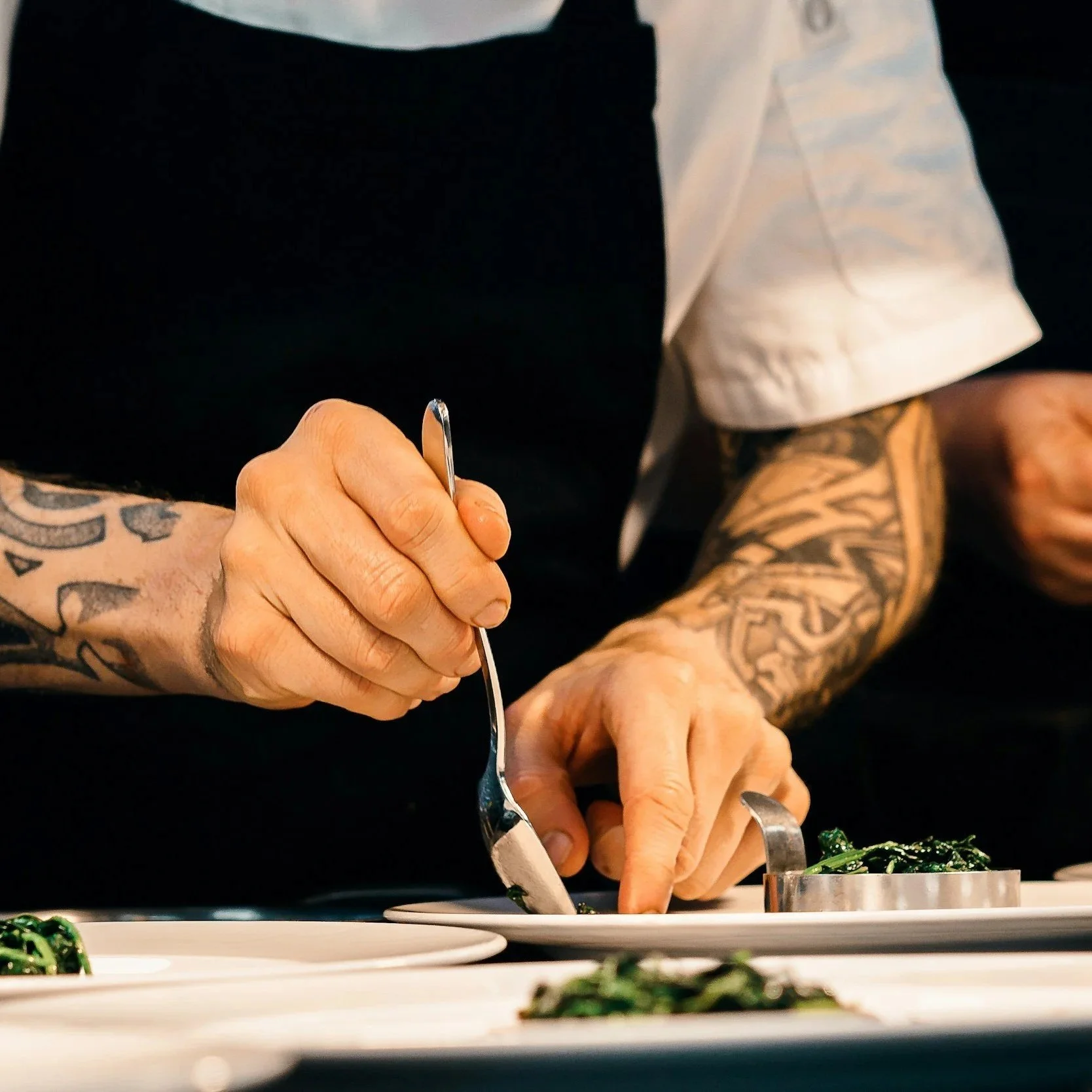 A chef with arm tattoos is preparing food, using kitchen tools, in a professional kitchen or restaurant setting.