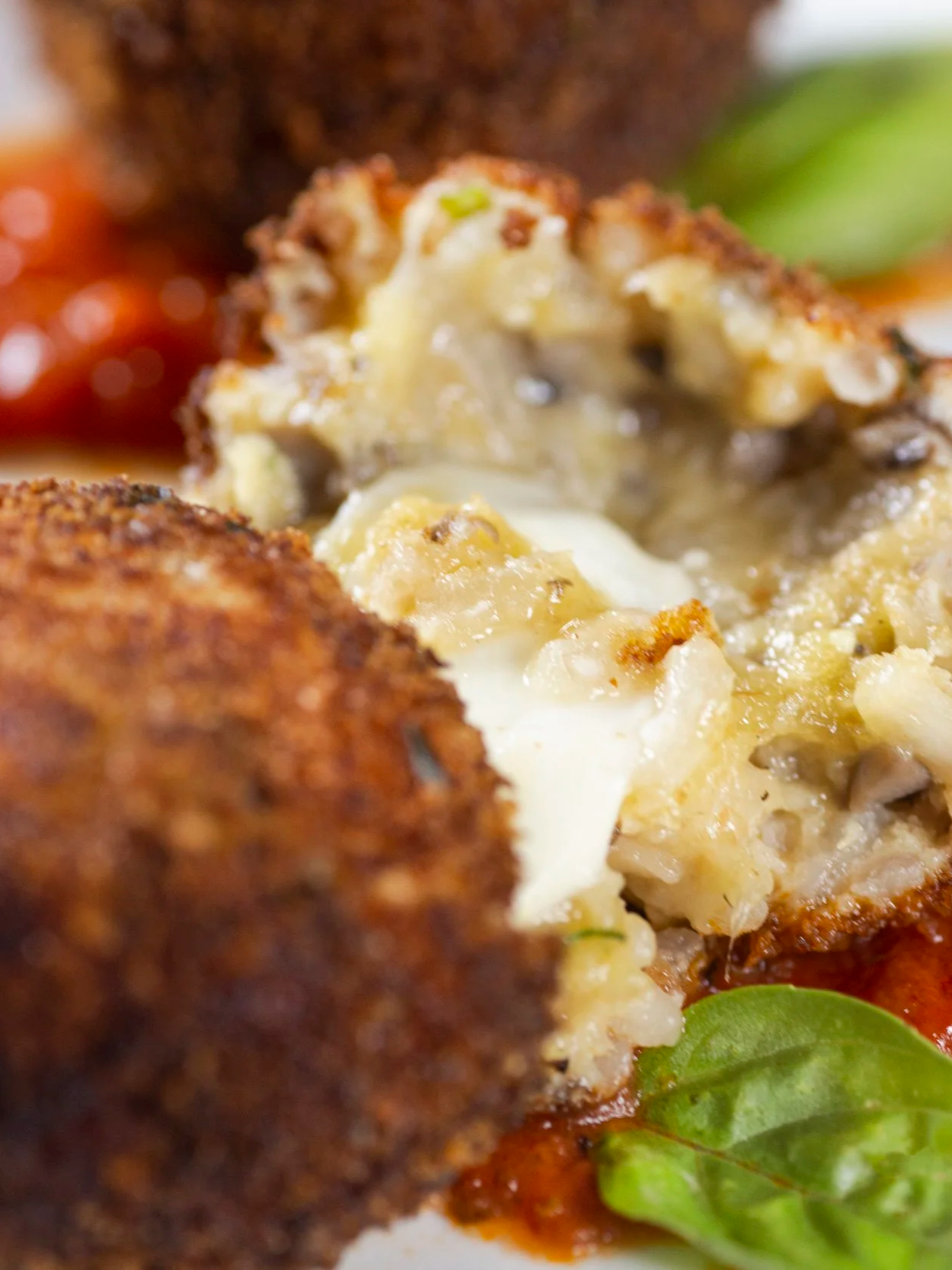 Close-up of a stuffed, breaded, and fried food item with creamy cheese and herbs inside, accompanied by some greens, tomatoes, and a red sauce.