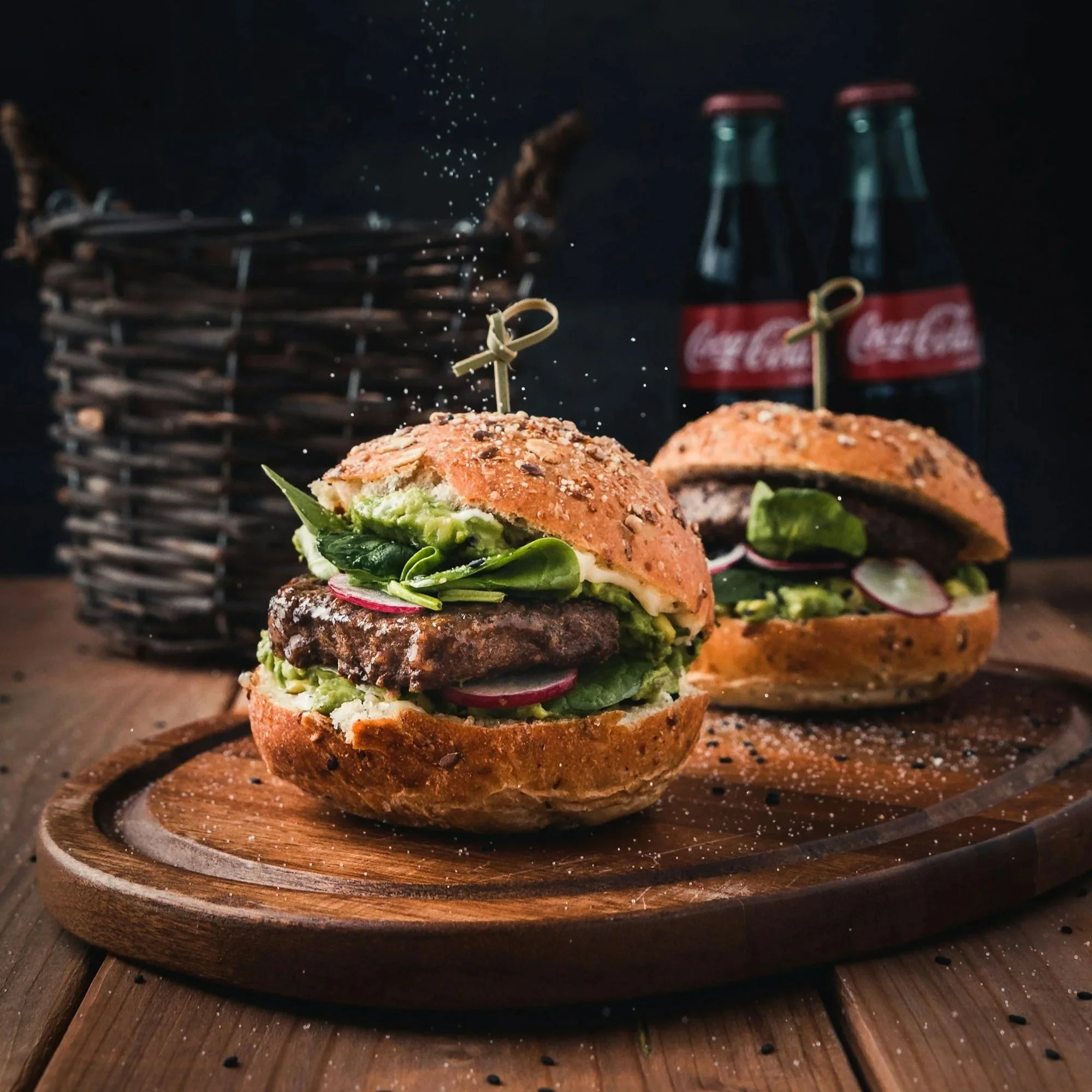 Close-up of spiced burger sliders on a wooden serving board, with a dark background and Coca-Cola bottles in the background.