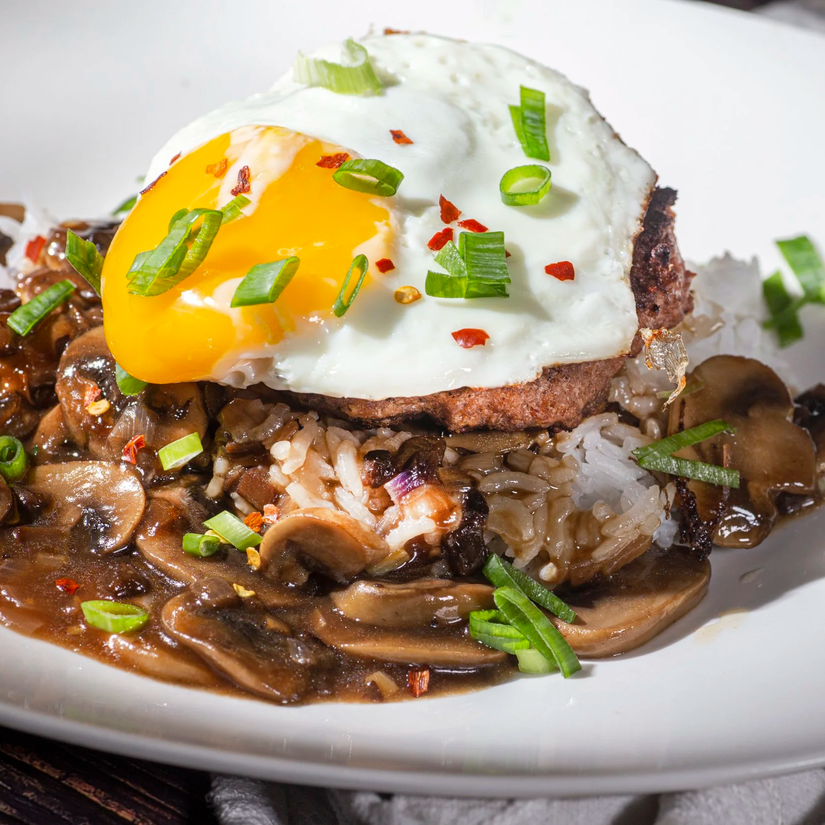 Close-up of a breakfast plate with a fried egg topped with green onions and red pepper flakes, served over sautéed mushrooms and rice with gravy.