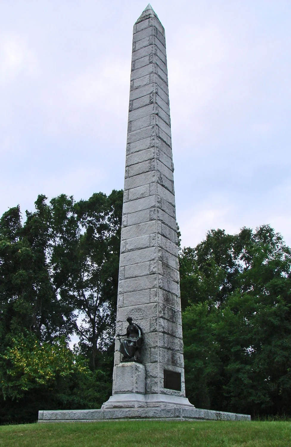A tall stone obelisk monument with a bronze statue of a seated figure at its base, set against green trees and a cloudy sky.